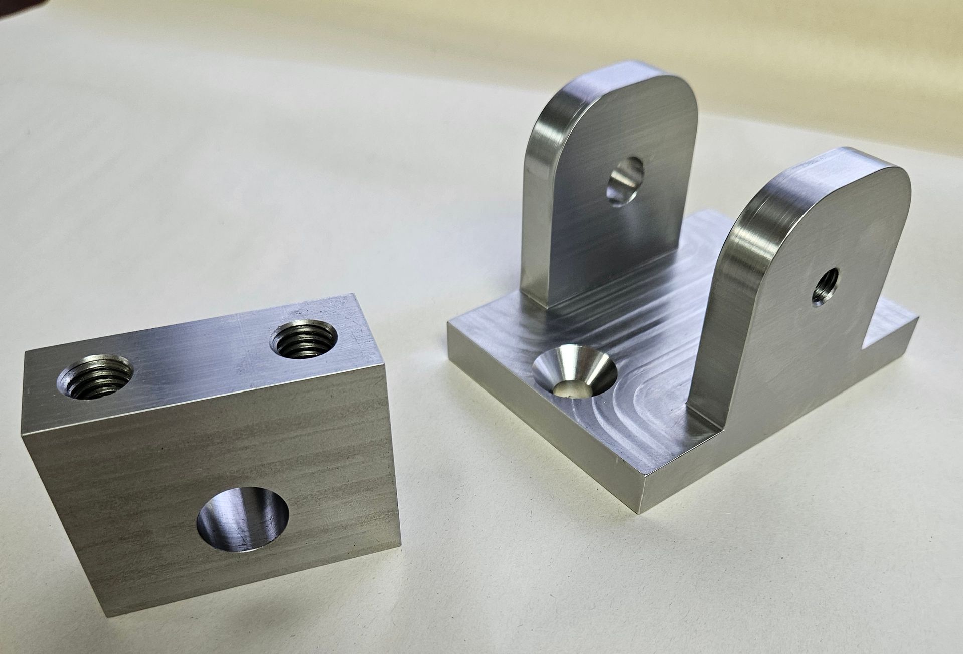 Two machined metal brackets sit on a plain surface, one U-shaped with holes and the other a block with threaded inserts.