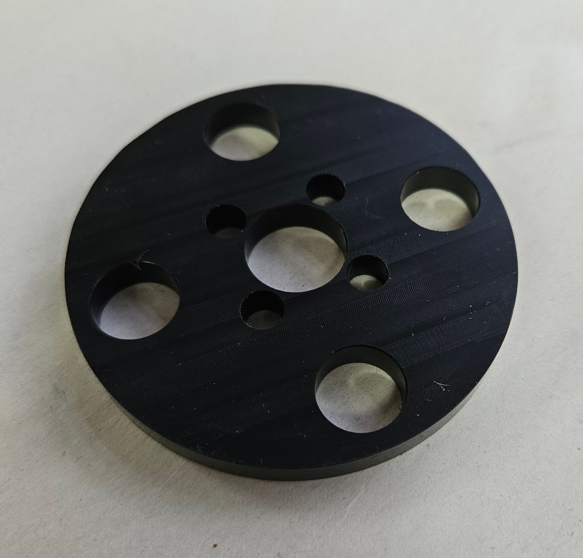 A flat, circular black metal mounting plate with a center hole and various smaller holes arranged in patterns.
