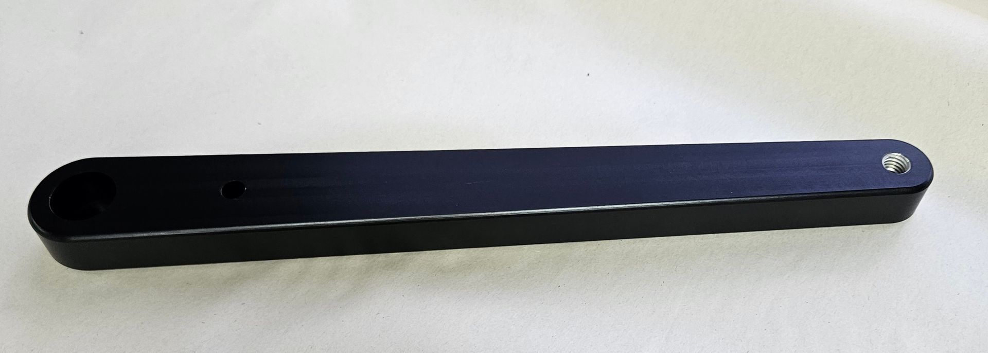 A black, elongated rectangular metal component with a rounded end featuring a circular hole, viewed against a light background.