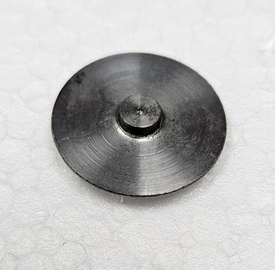 A small, dark grey, circular metal component with a raised, cylindrical center sits on a textured white surface.