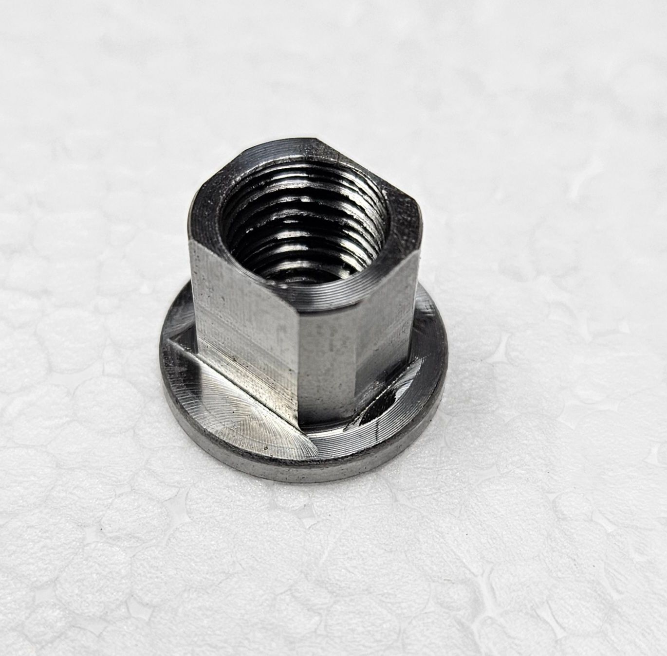 A silver-colored flanged hex nut with visible internal threads, resting on a white, textured surface.