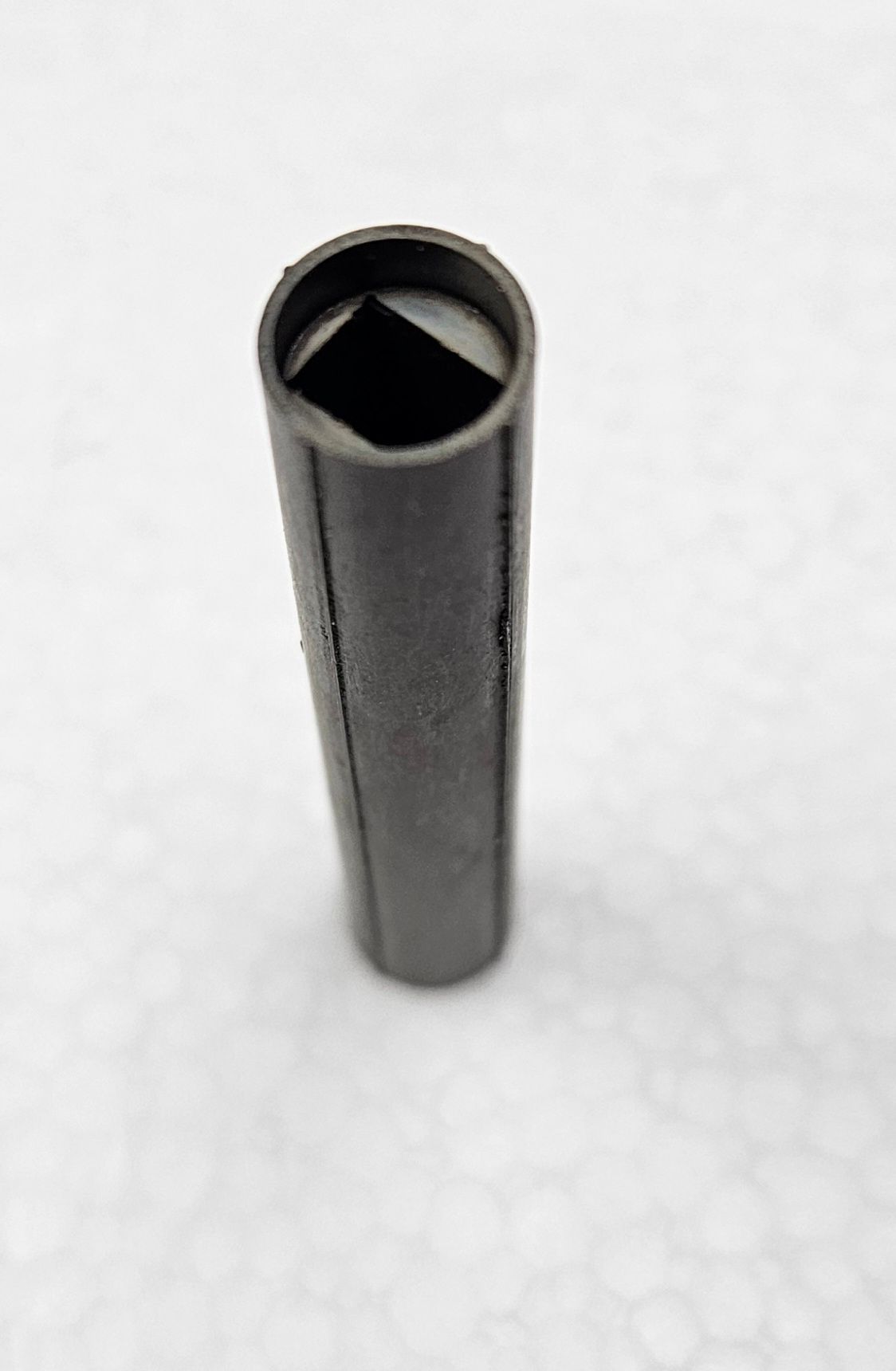 A dark gray, cylindrical tool with a square-shaped hollow opening at the top, set against a white, textured background.
