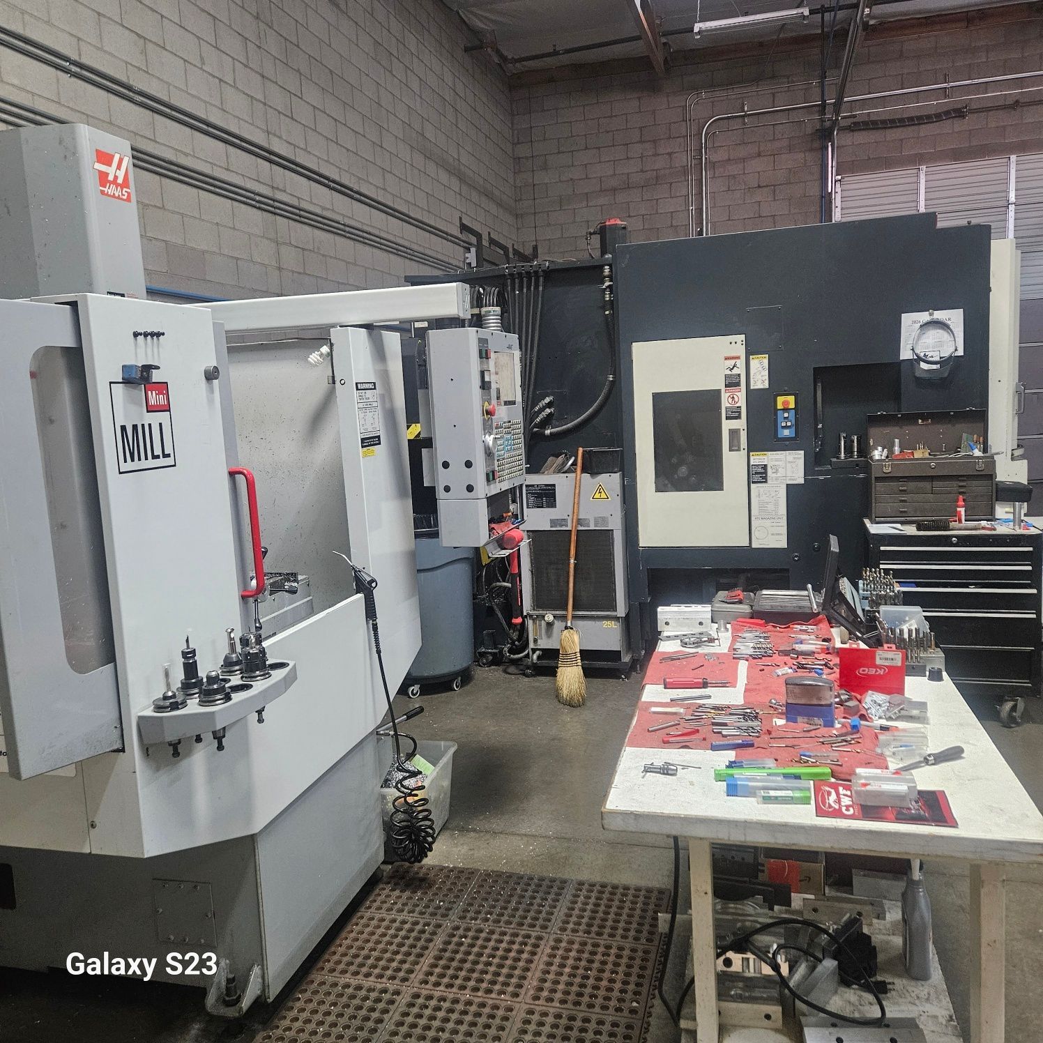 A machine shop featuring two industrial CNC milling machines and a workbench cluttered with tools and metal components.