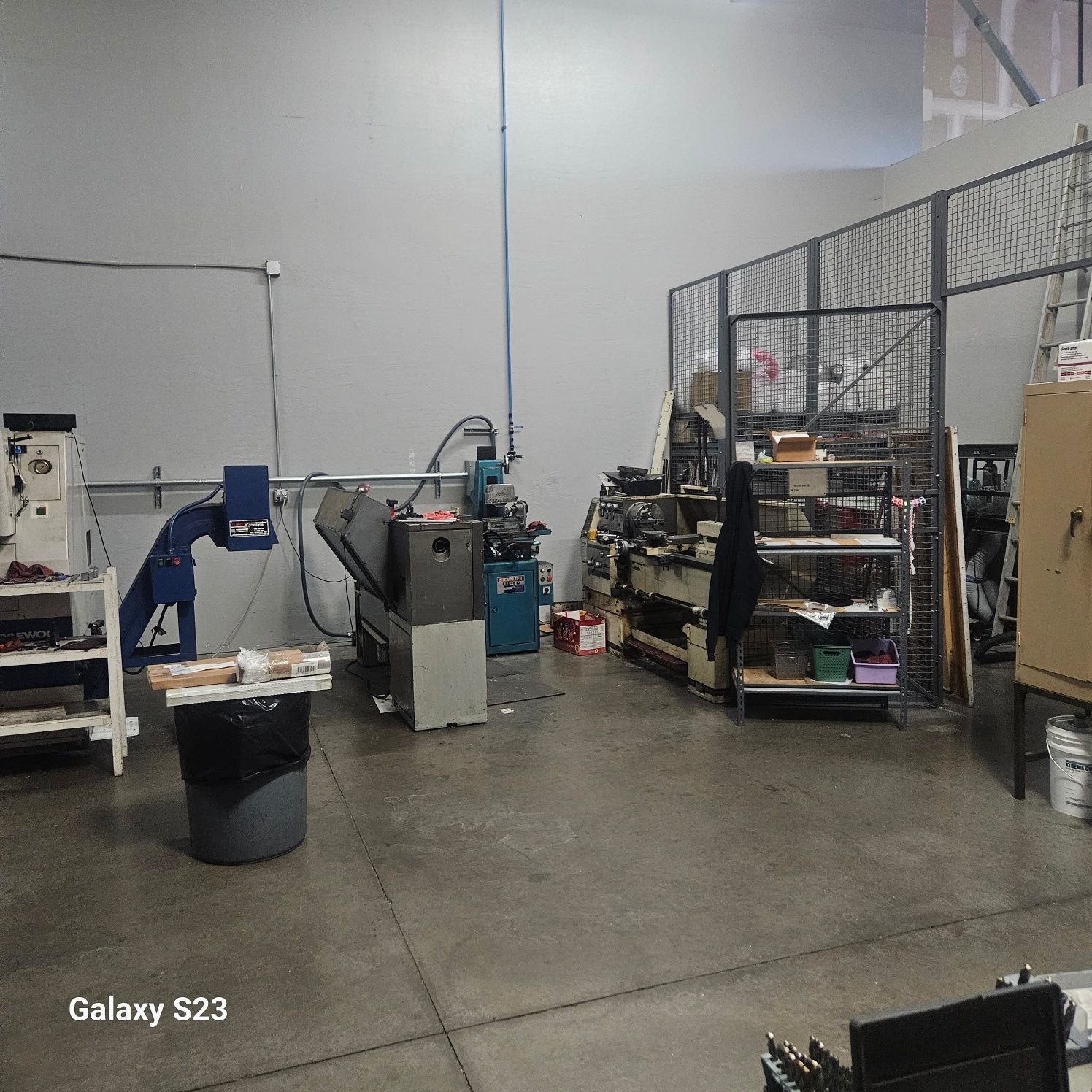 A workshop featuring metalworking machinery, shelves with tools, a metal cage enclosure, and a gray concrete floor.