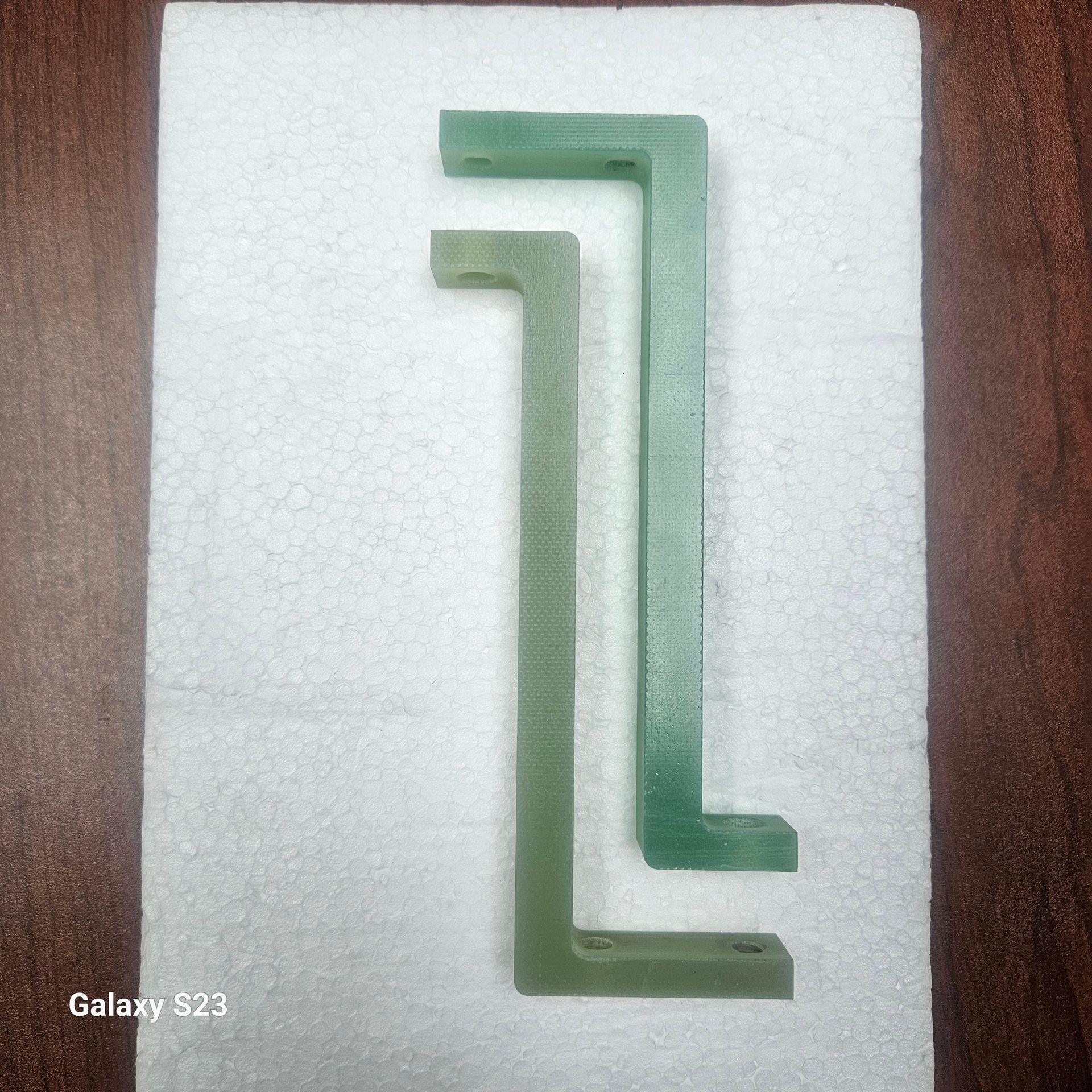 Two L-shaped, light green fiberglass brackets laid side-by-side on a white styrofoam surface.