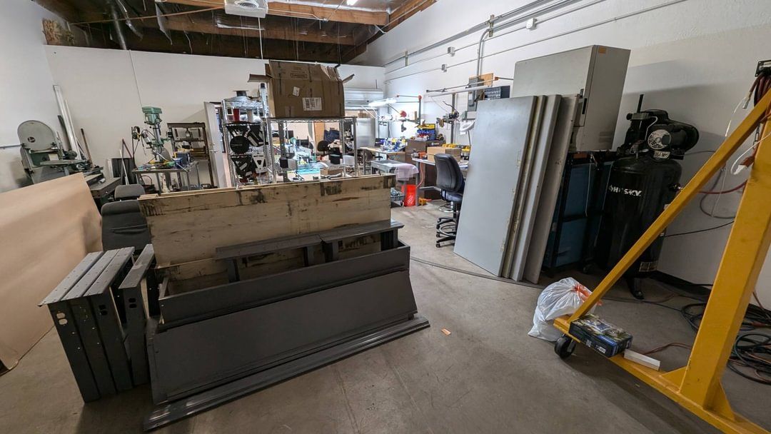 A cluttered workshop featuring metal workbenches, large gray panels, tools, and a yellow gantry crane.