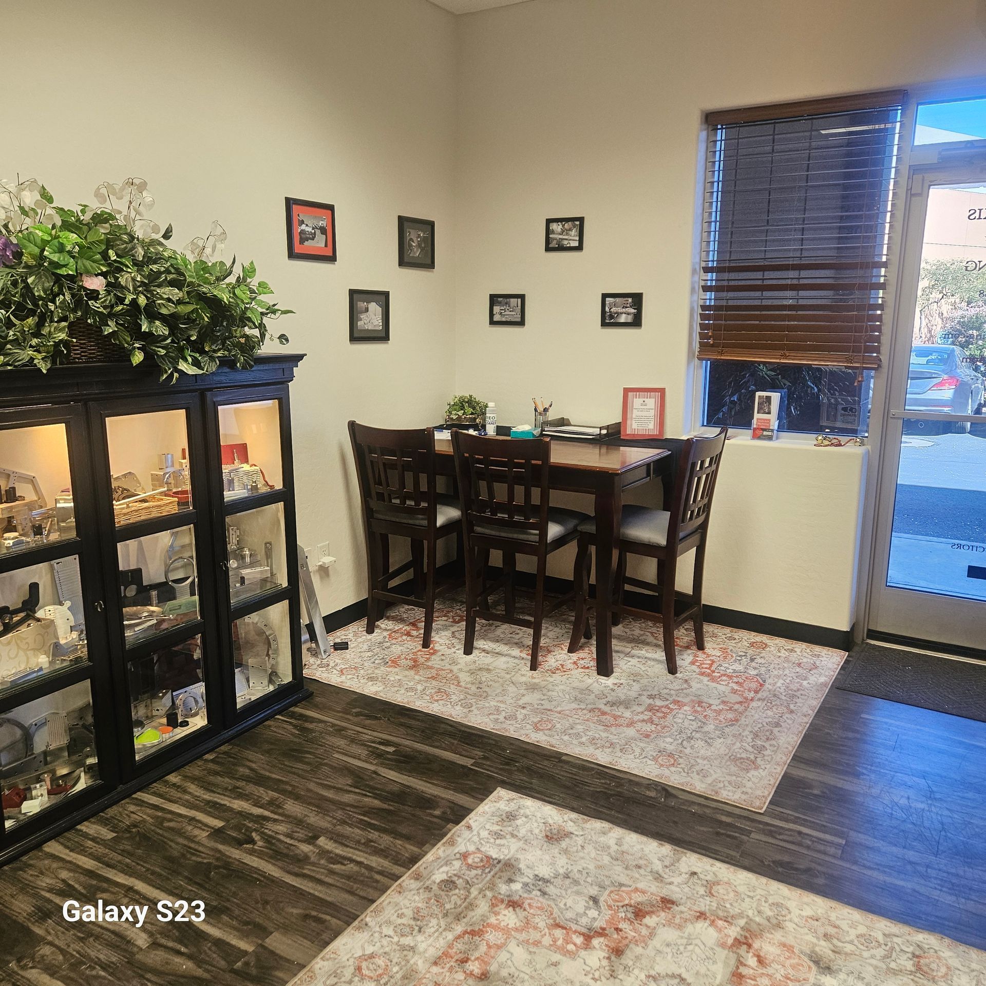 A room with dark wood flooring, a decorative display cabinet, a dining set with three chairs, and wall-mounted photos.