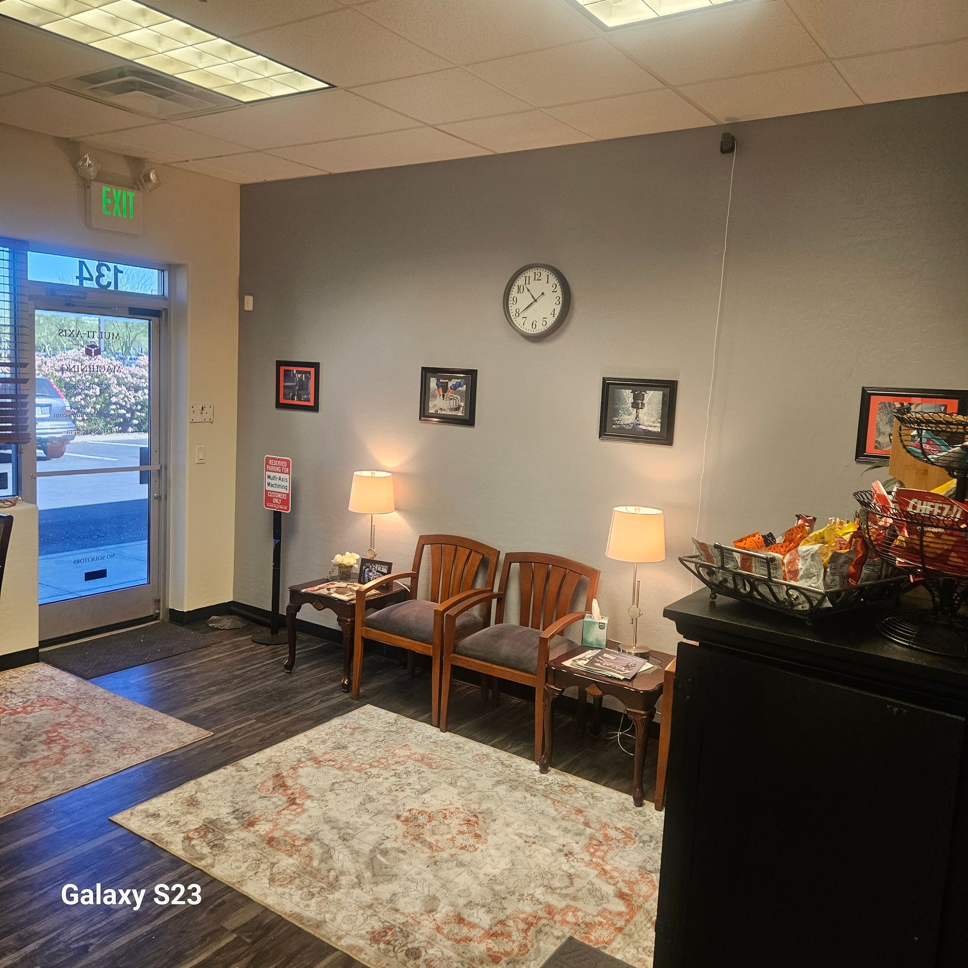 A waiting room with two wooden chairs, lamps, wall art, a clock, and a snack display on a black cabinet.