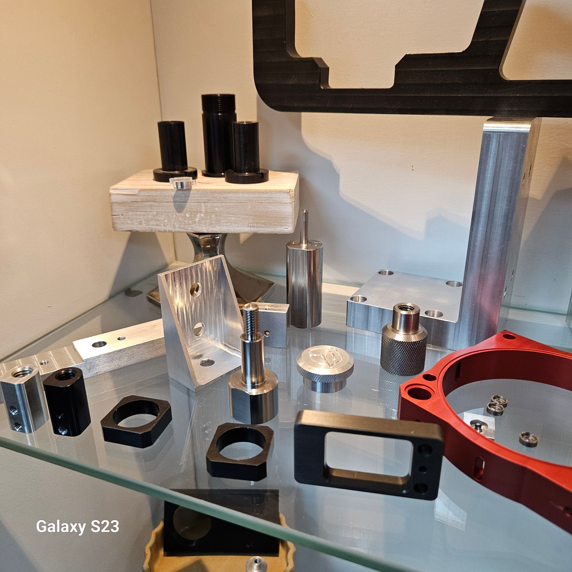A collection of machined metal and plastic mechanical parts, including brackets and cylinders, arranged on a glass shelf.