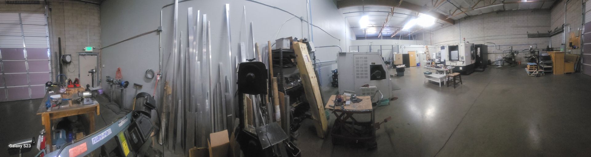 A wide-angle panoramic view of an industrial machine shop with raw metal materials, workbenches, and large equipment.