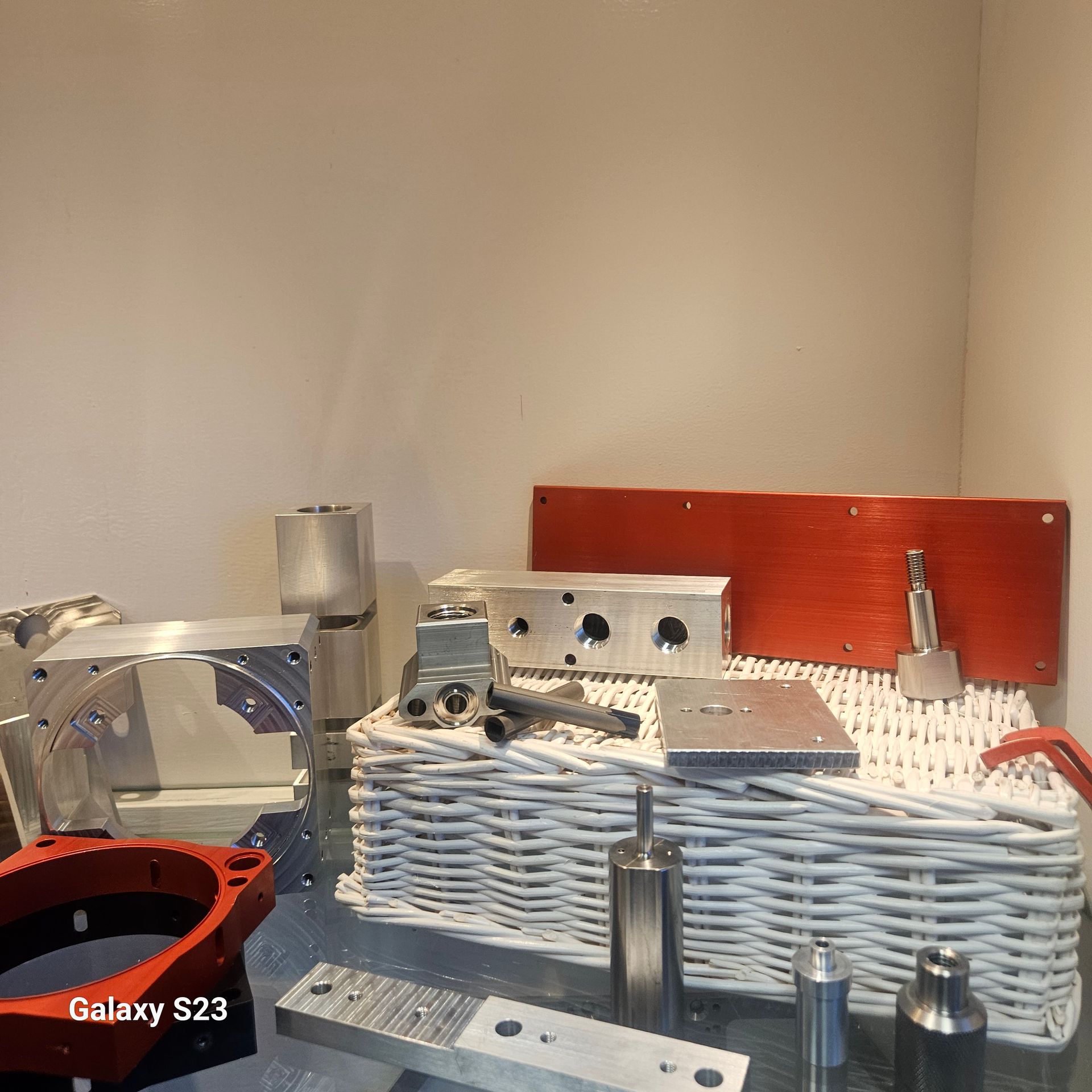 A collection of various machined metal components, including aluminum blocks and red plates, resting on a white basket.