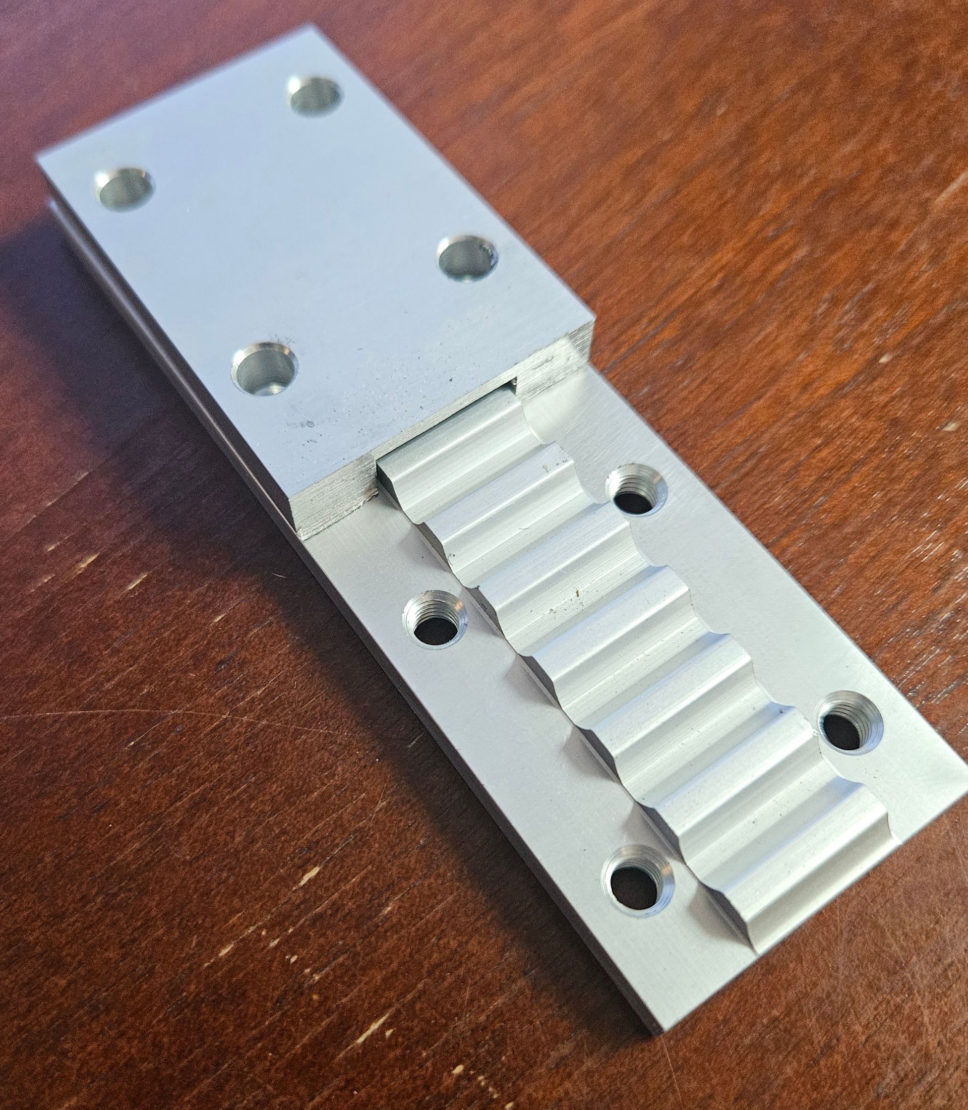 A rectangular aluminum bracket with a flat mounting area containing four holes and a serrated, gear-like central section.