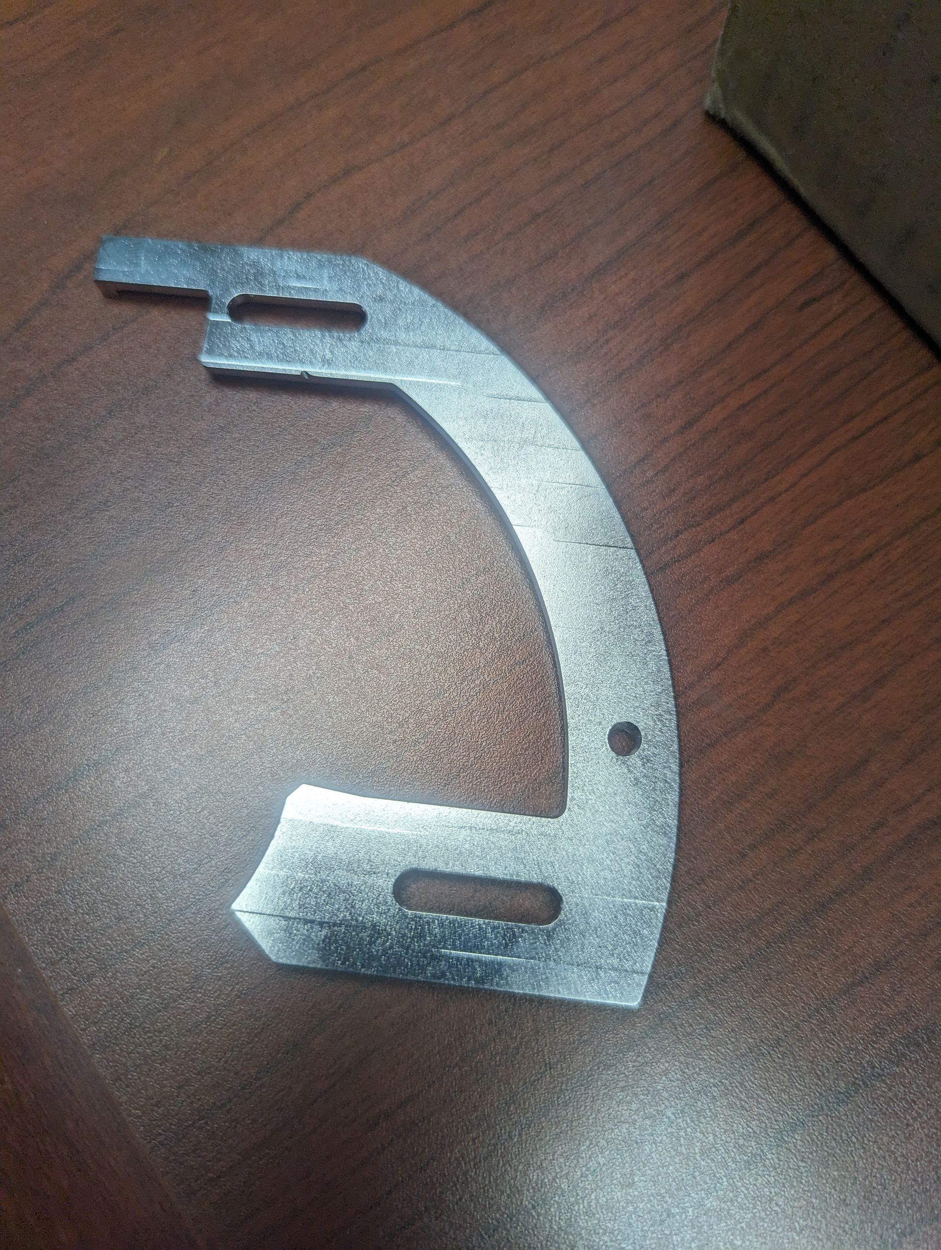 A curved, silver metal bracket with two elongated slots and one small hole, lying on a dark wooden surface.