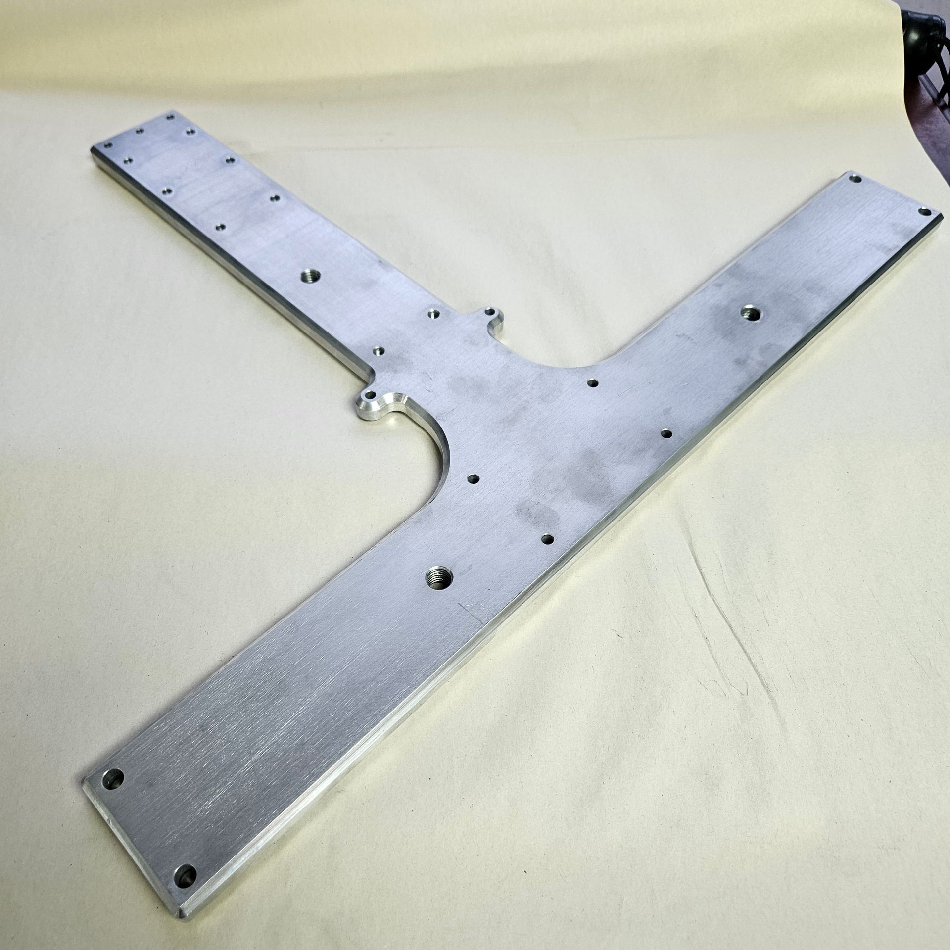 A T-shaped, machined aluminum plate with multiple drilled holes and mounting points on a light-colored surface.