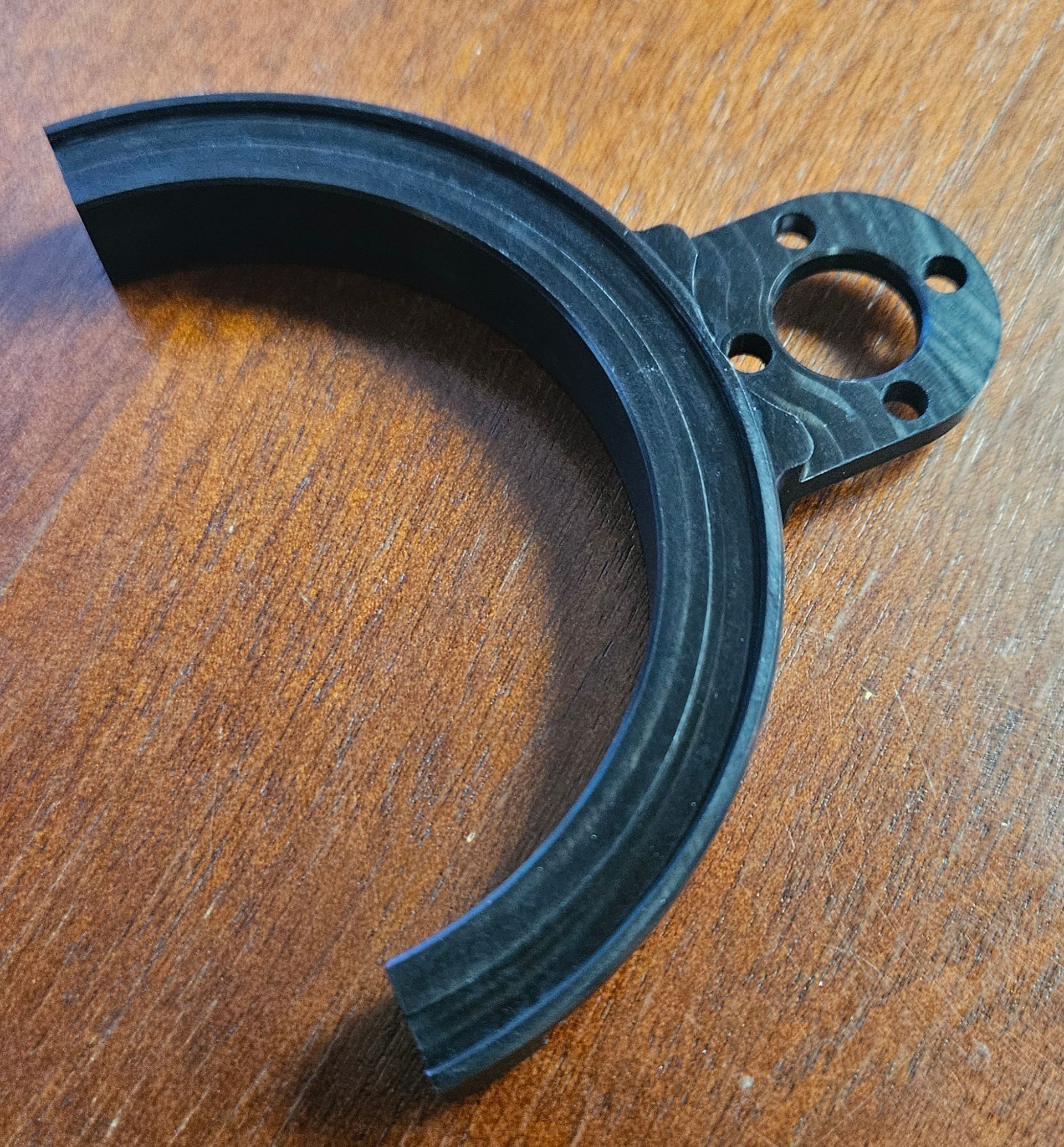 A black, 3D-printed curved bracket with a circular, multi-holed mounting tab, resting on a wooden surface.