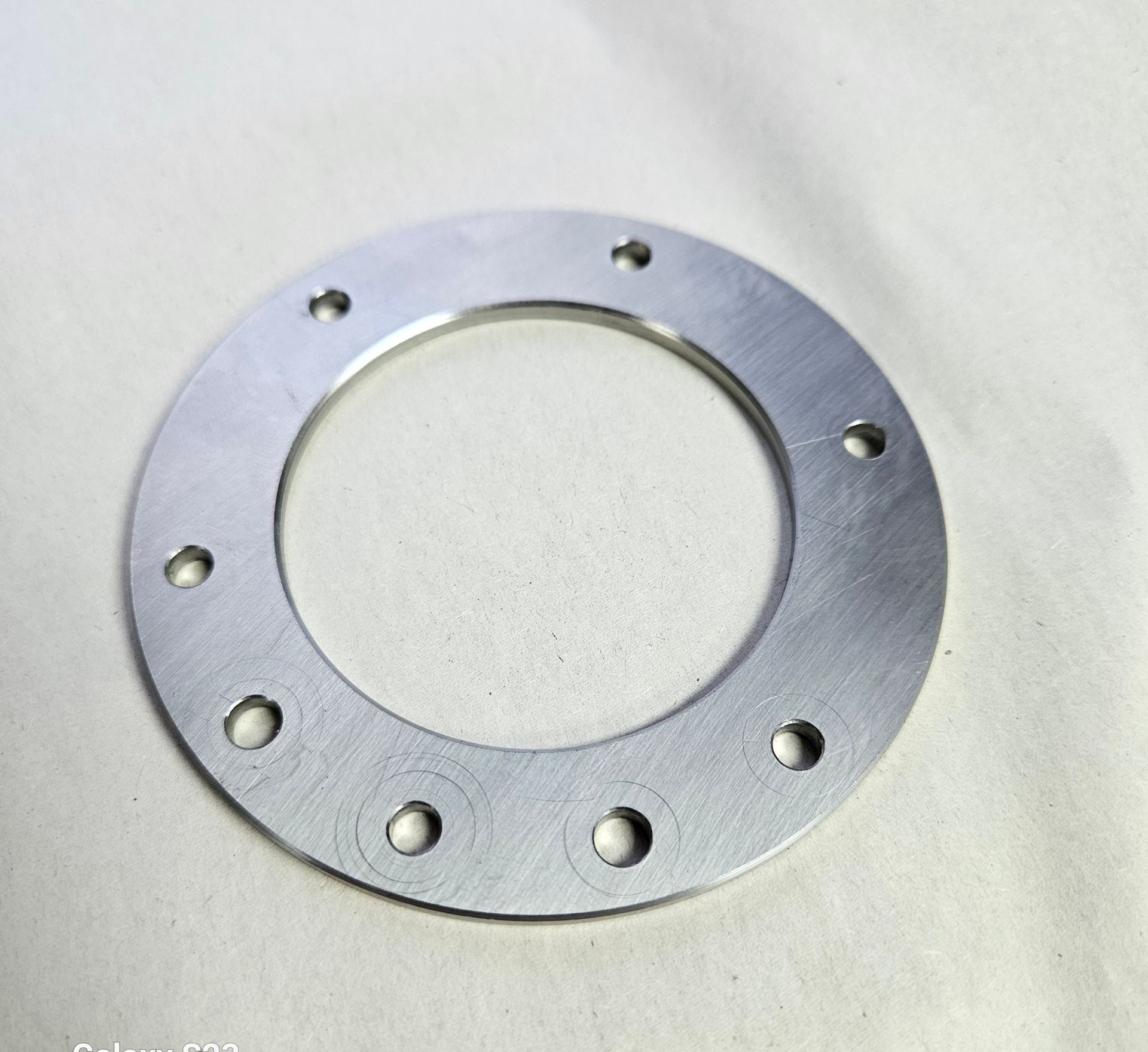 A flat, circular metal washer with eight evenly spaced holes around its perimeter, resting on a white fabric surface.