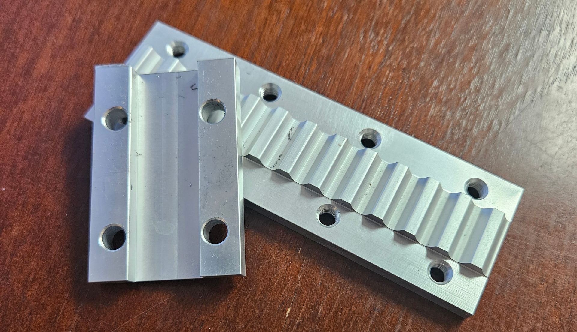 Two machined aluminum metal plates with grooved channels and mounting holes, resting on a dark wooden surface.