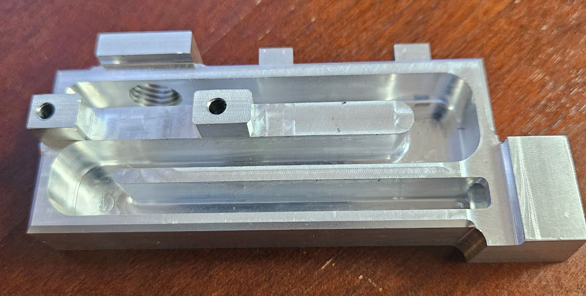 A small, silver-colored, machined metal part with intricate internal channels, holes, and a threaded feature.