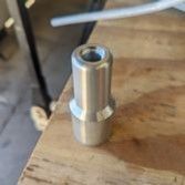 A silver-colored, machined metal cylindrical adapter or bushing stands upright on a wooden workbench.