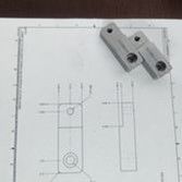 Two identical, small gray metal components rest on a technical drawing showing top and side view schematics.