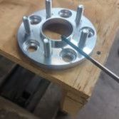 A silver metal wheel spacer with five integrated studs sitting on a wooden surface, being touched by a metal tool.