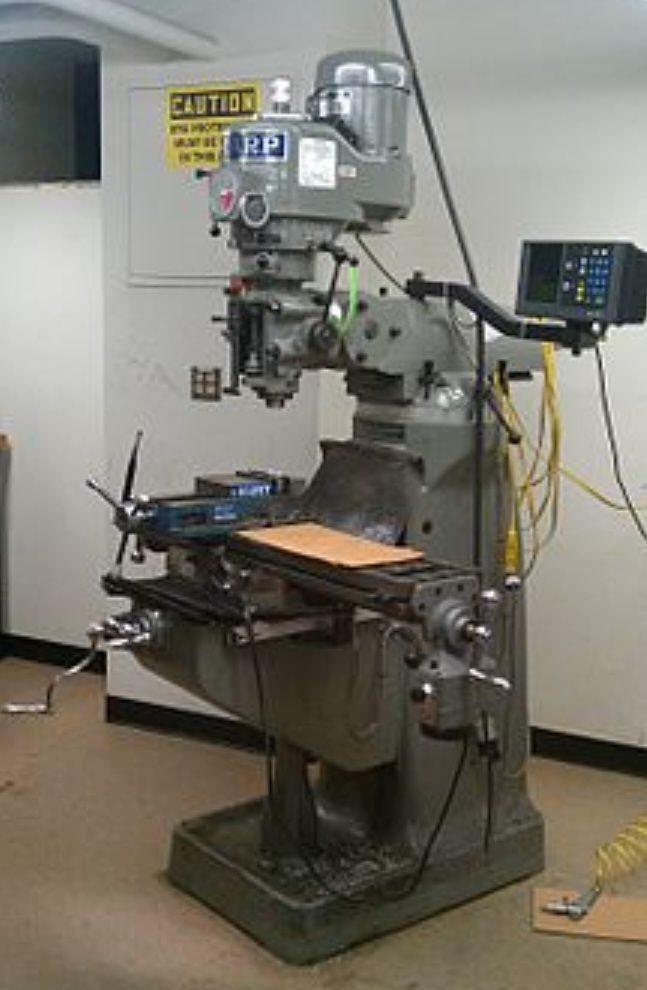 A gray industrial milling machine stands in a workshop with a digital readout display arm and a cautionary wall sign.