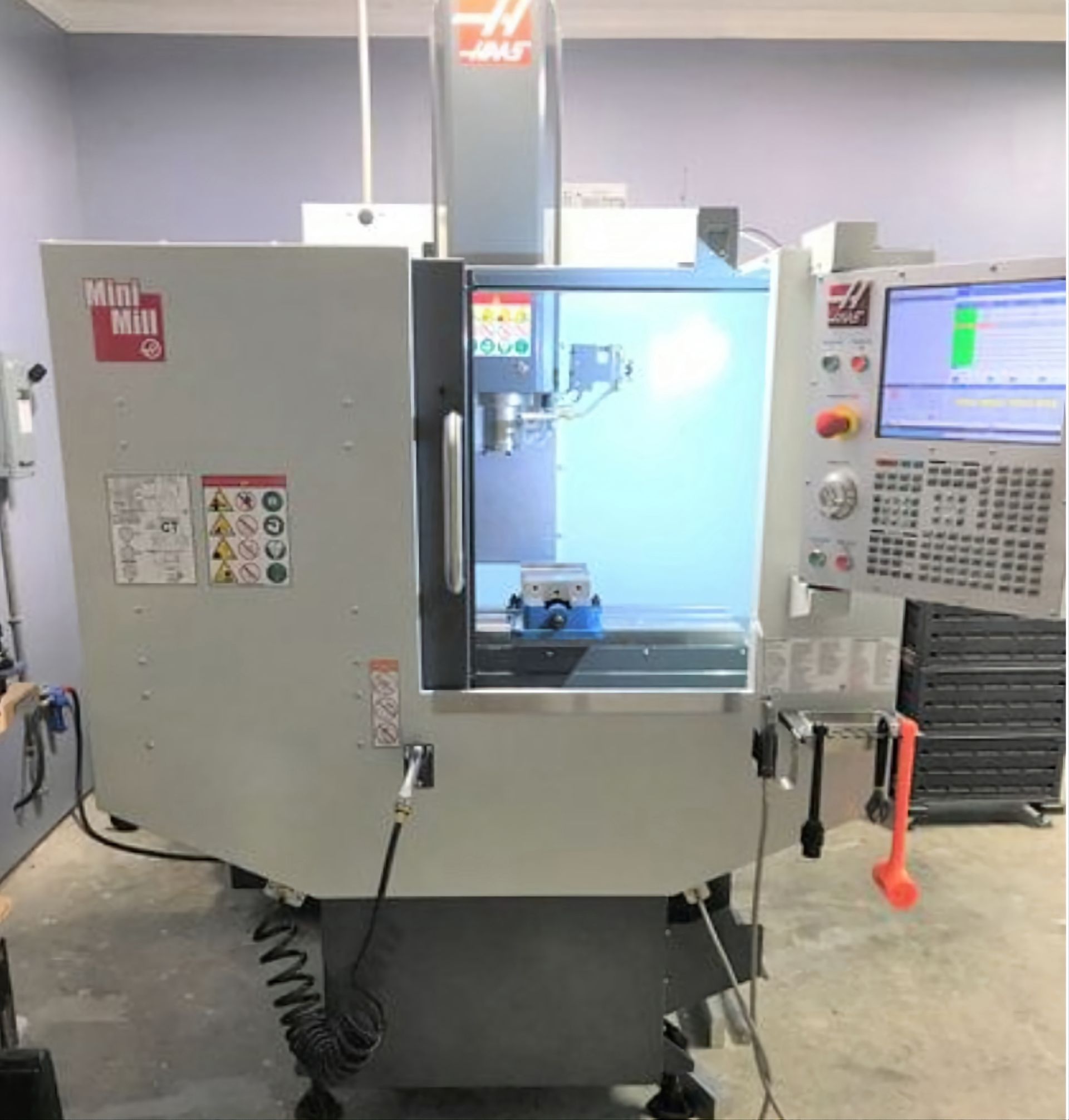 A gray Haas Mini Mill CNC machine stands in a workshop, featuring a clear access door, a screen, and a control panel.
