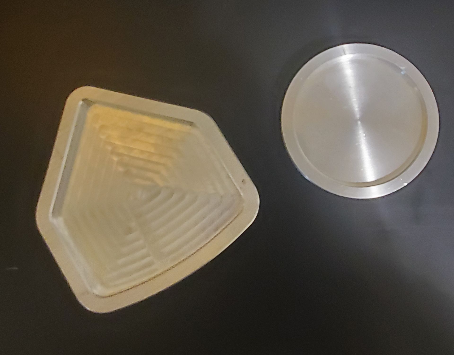 A metallic, pentagon-shaped container with ridged interior lines rests beside a circular, smooth metal lid on a dark surface.