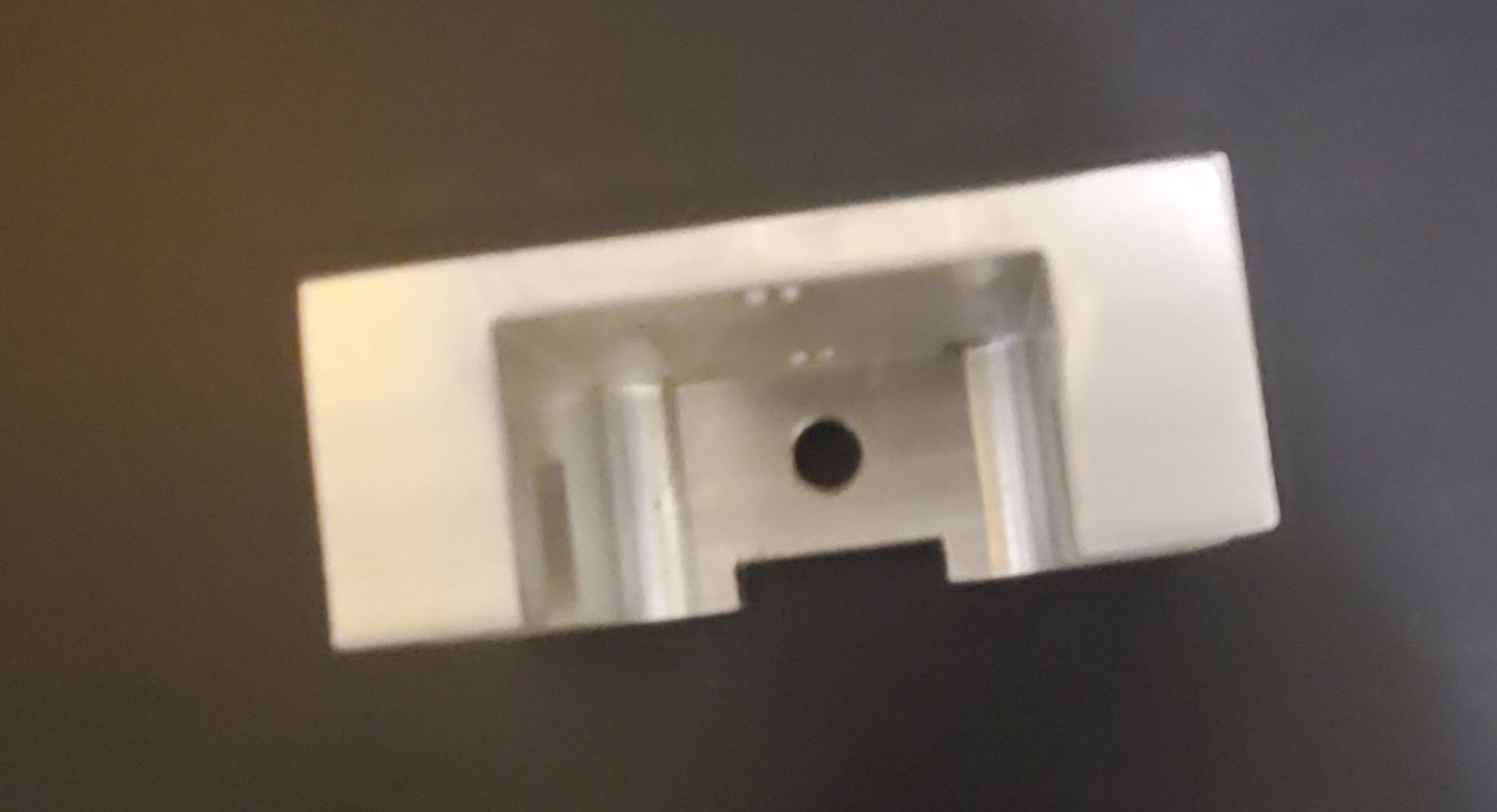 A light grey, rectangular metal block with a hollowed-out center, mounting holes, and recessed interior features.