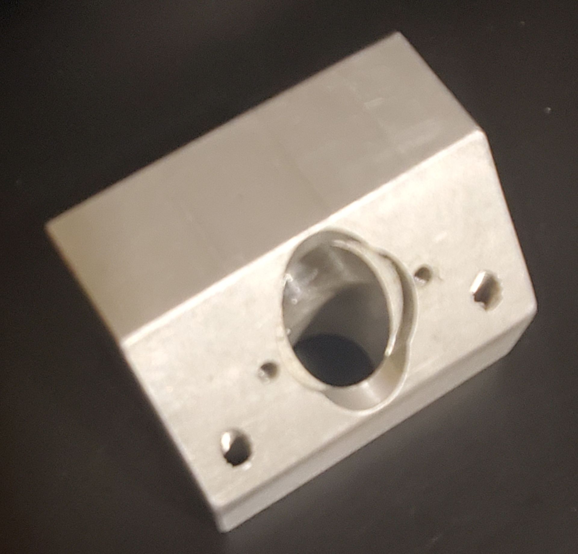 A silver, wedge-shaped metal bracket with a central circular hole and four mounting holes, set against a dark background.