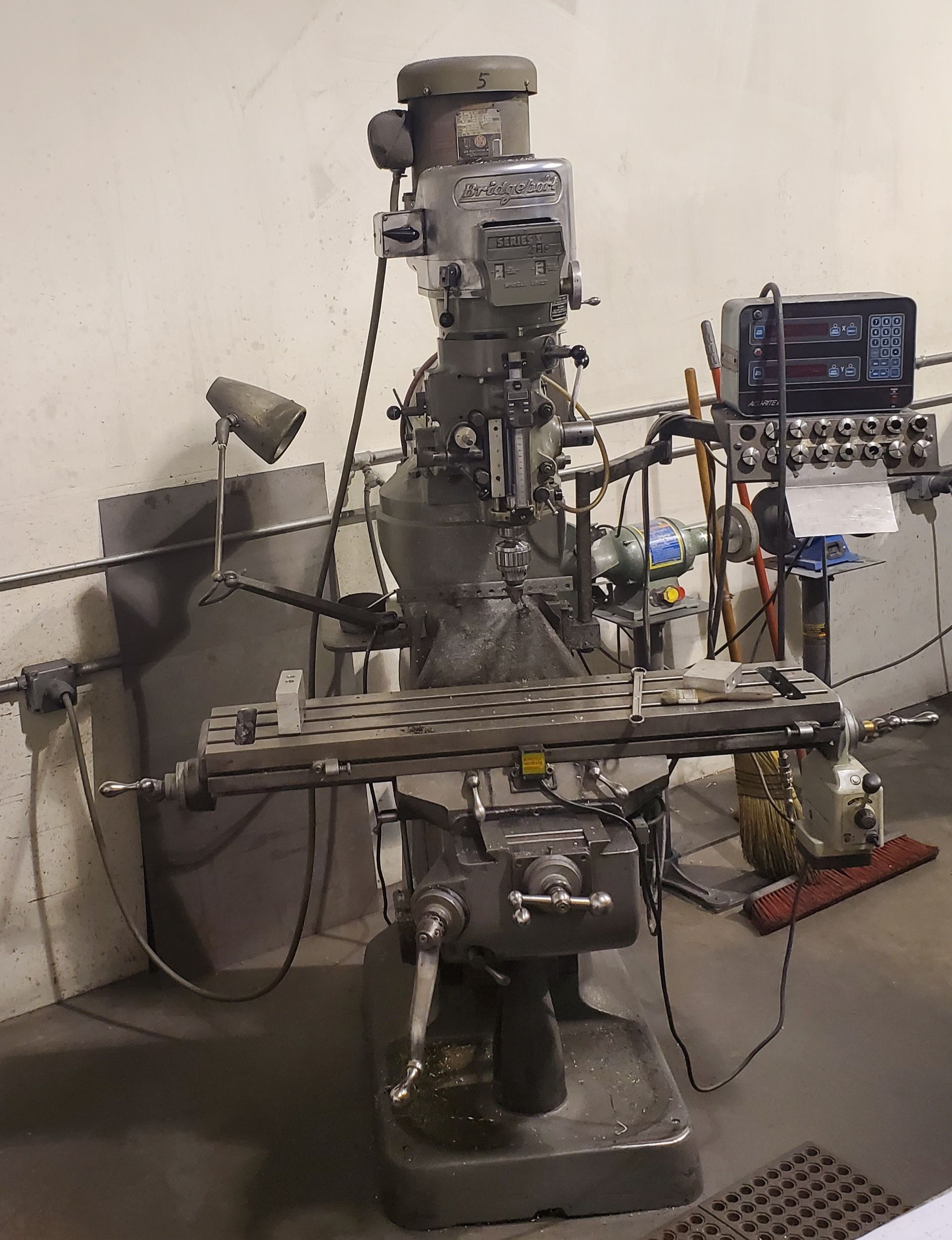 A gray vertical milling machine sits in a workshop, featuring a work table, a spindle head, and a digital control box.