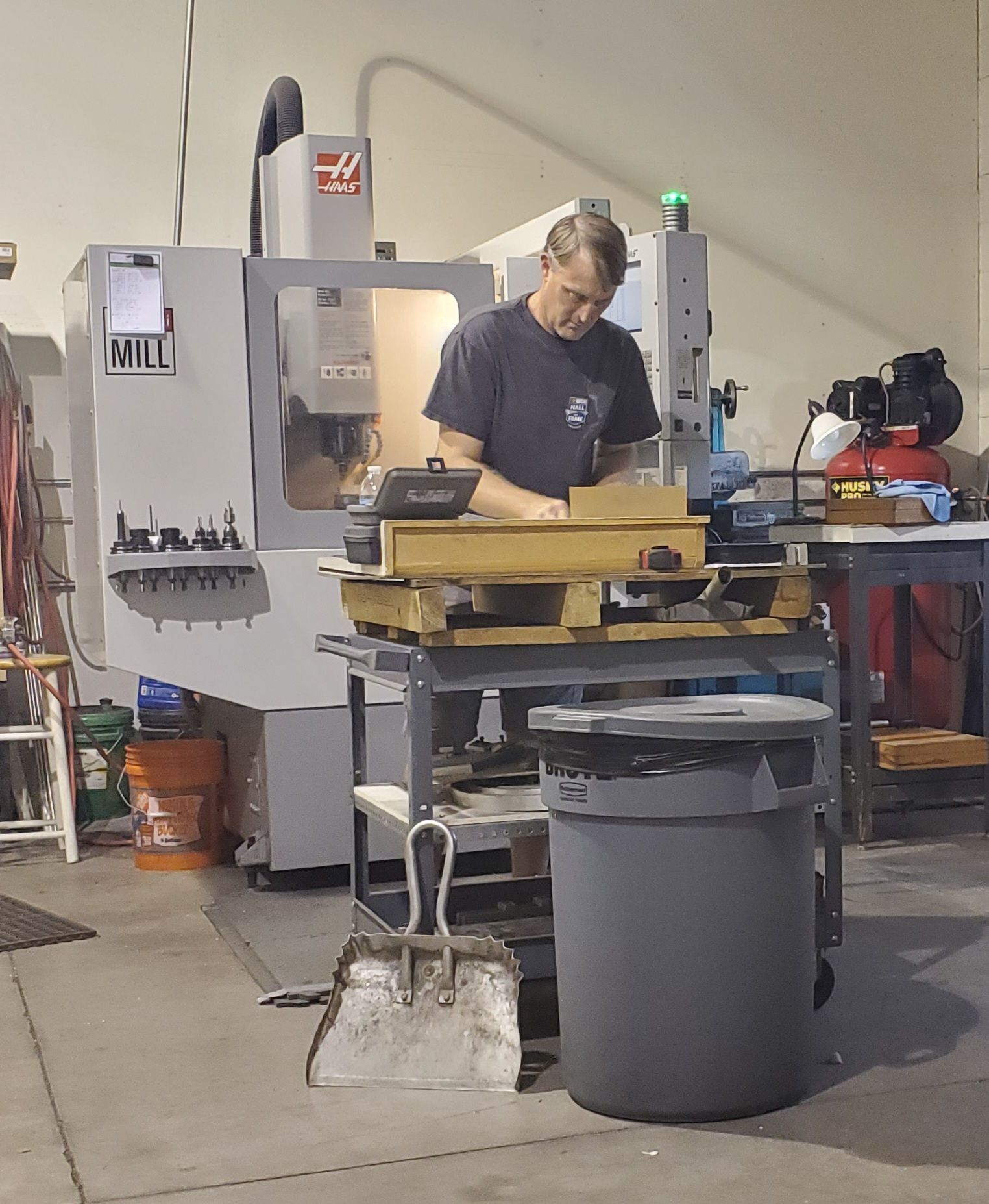 A person works at a metal-working machine in a factory, standing at a workbench with a trash can in the foreground.