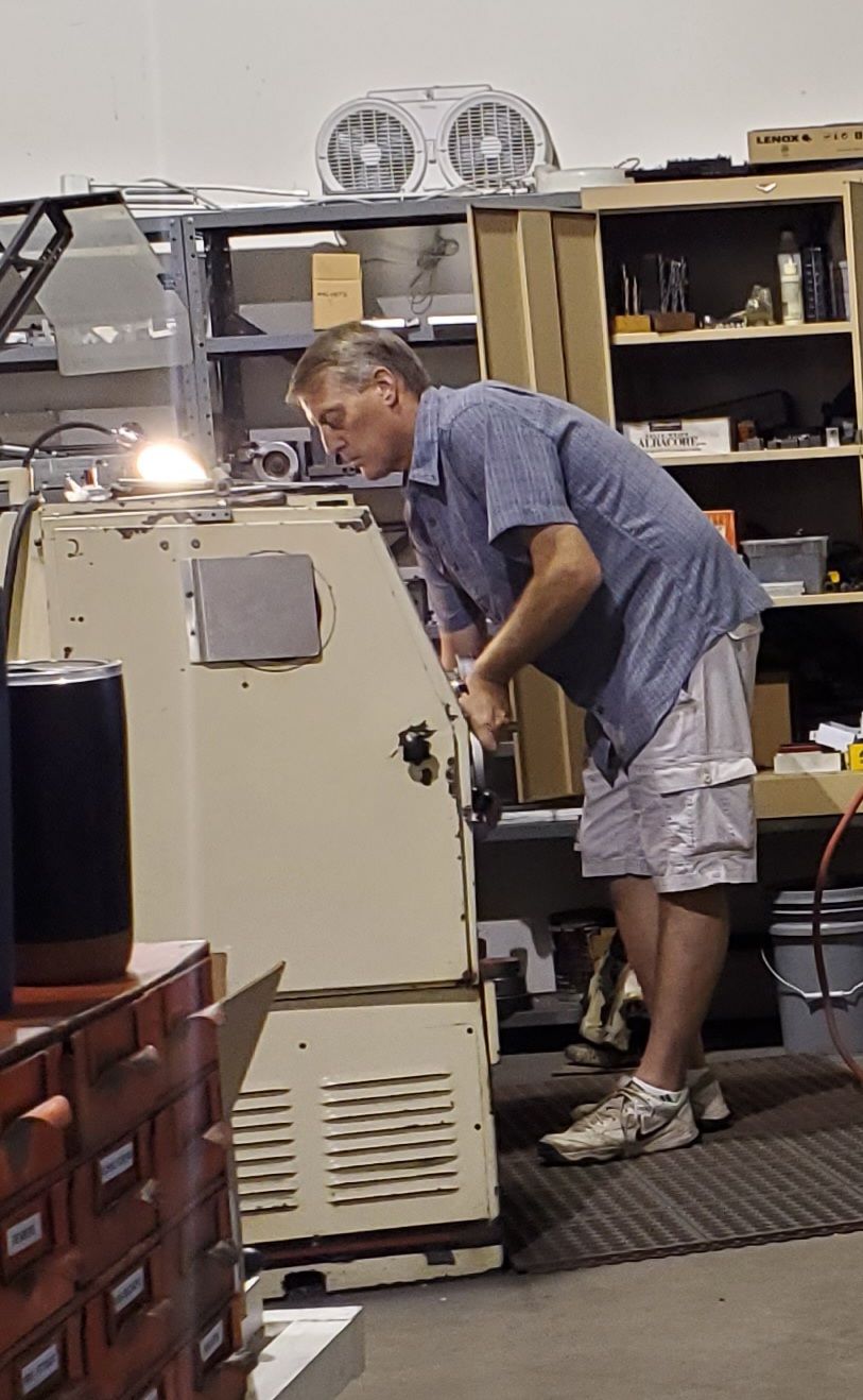 A person in a blue shirt and shorts stands in a workshop, working intently on a piece of machinery.