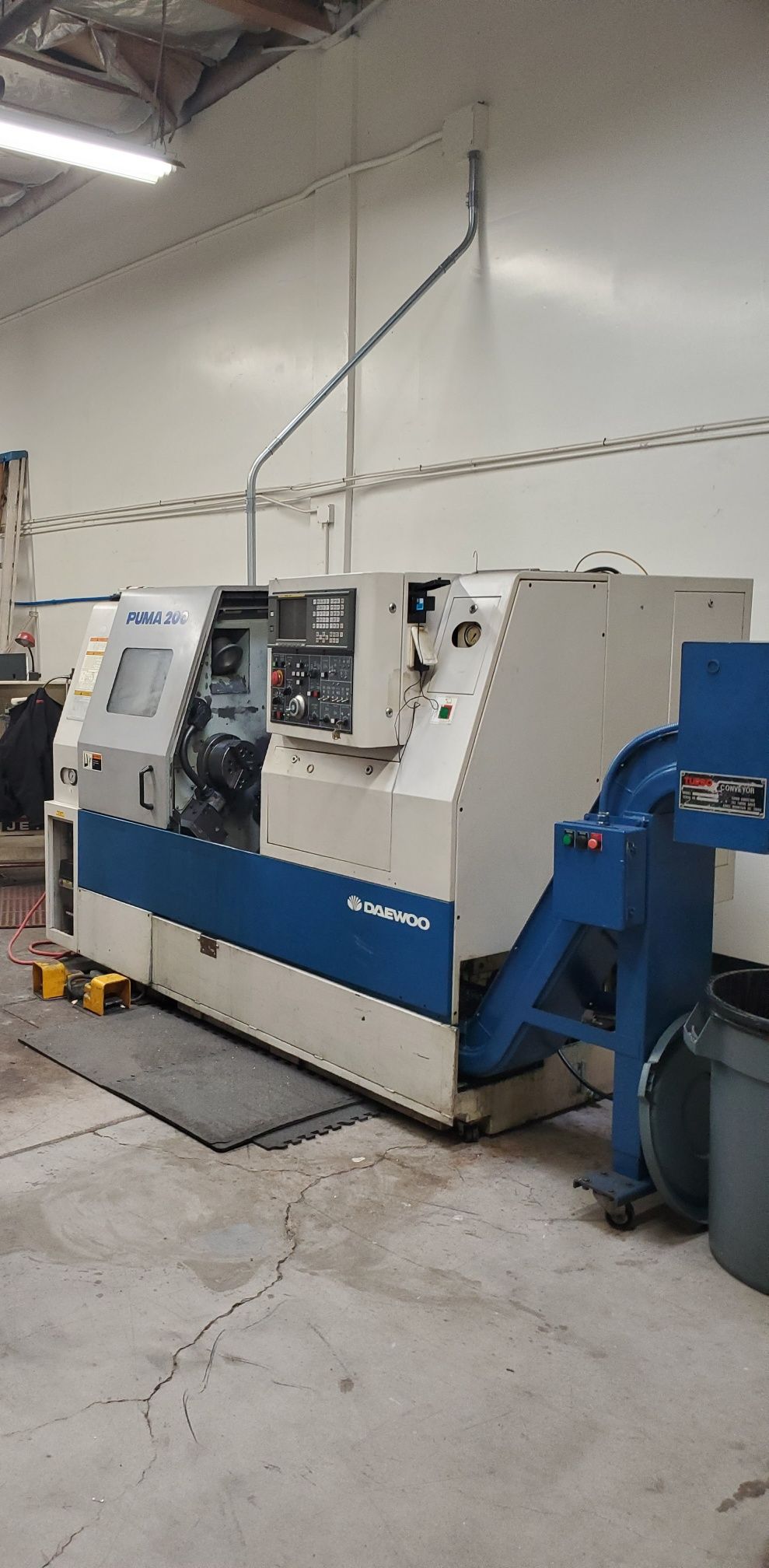 A white and blue CNC lathe sits on a concrete floor in a shop with a tall ceiling and overhead lighting.