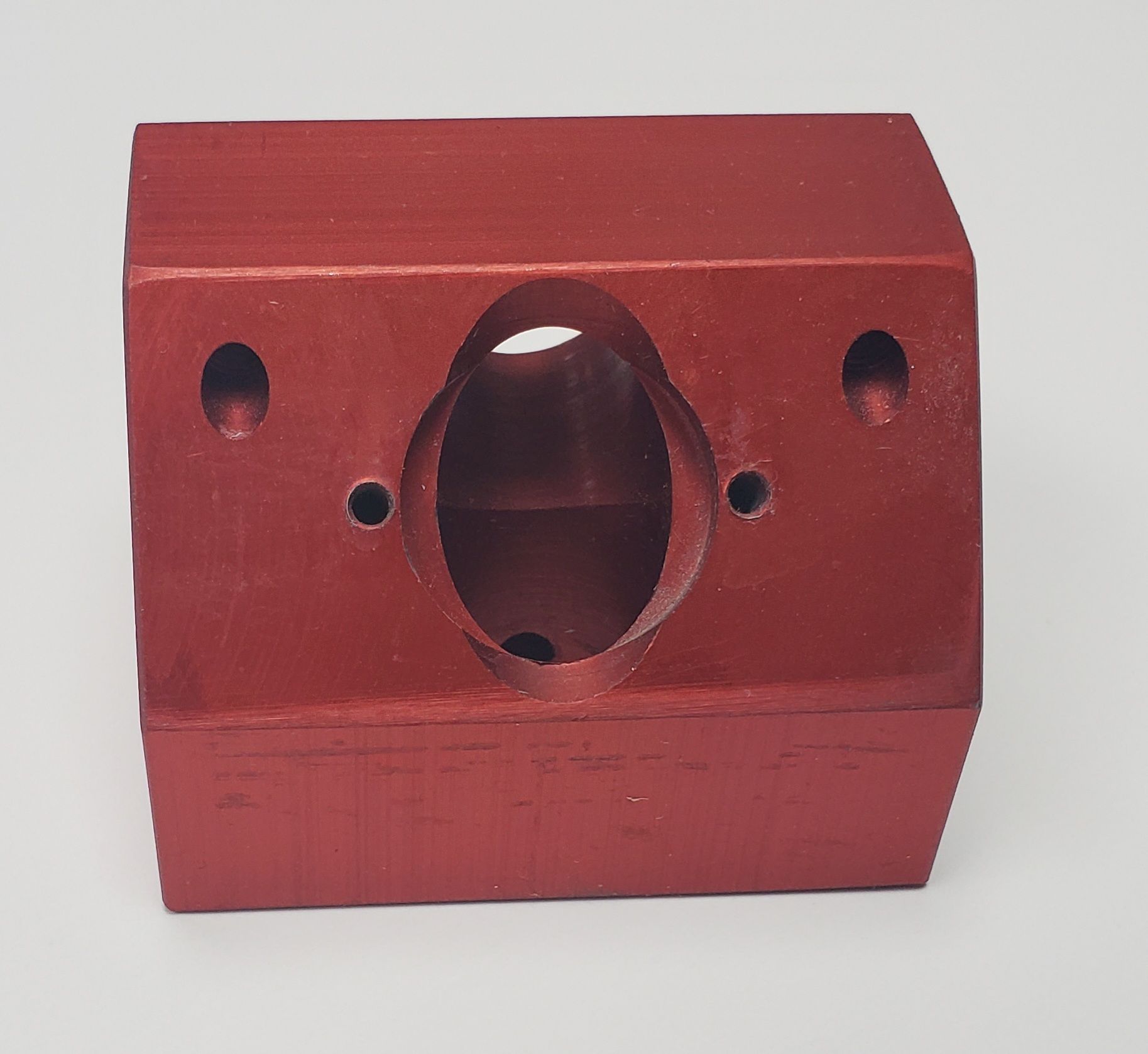A red, machined metal block with a central circular opening and four smaller mounting holes on its face.