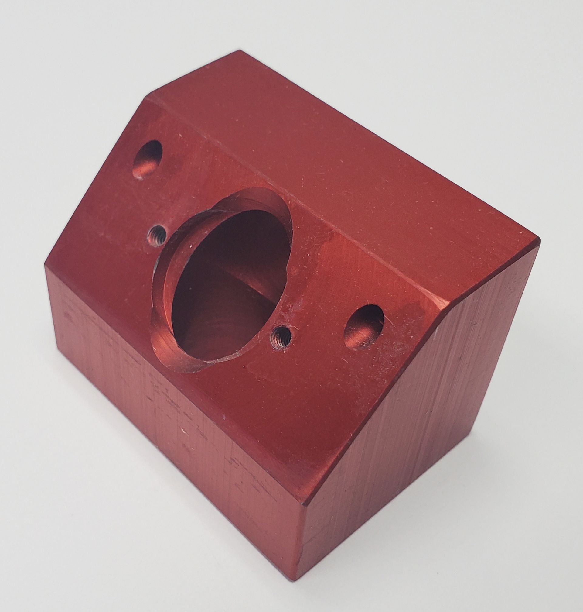 A red, angled metal block featuring a central circular opening surrounded by four smaller mounting holes.