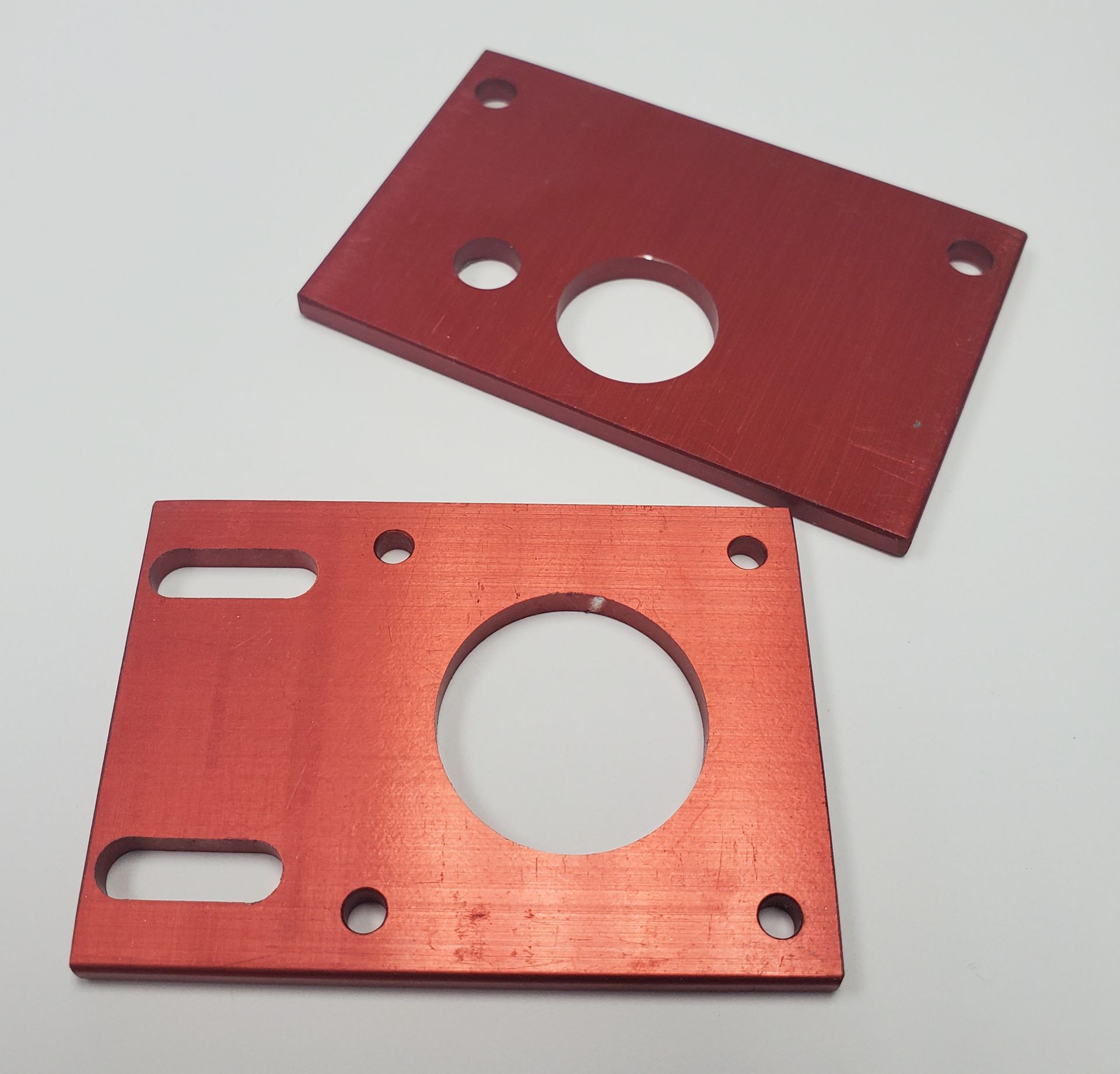 Two red rectangular metal plates with circular and slotted cutouts lie against a plain white background.