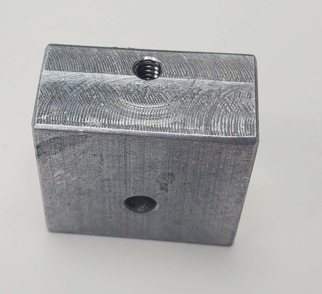 A small, rectangular gray metal block with a threaded hole on the top edge and a circular hole on its front face.