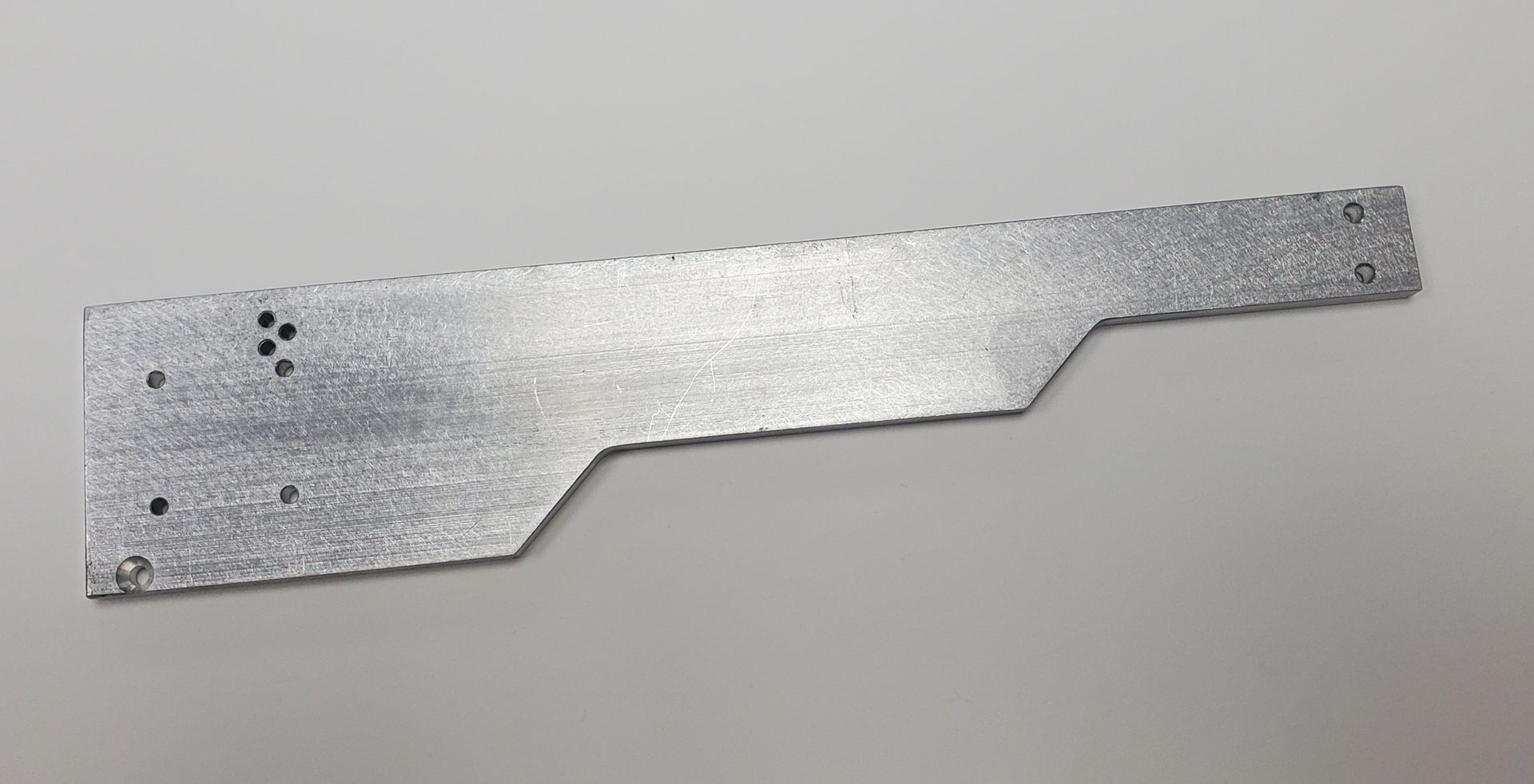 A flat, silver-colored metal plate with a stepped profile and several small drilled holes on a light background.