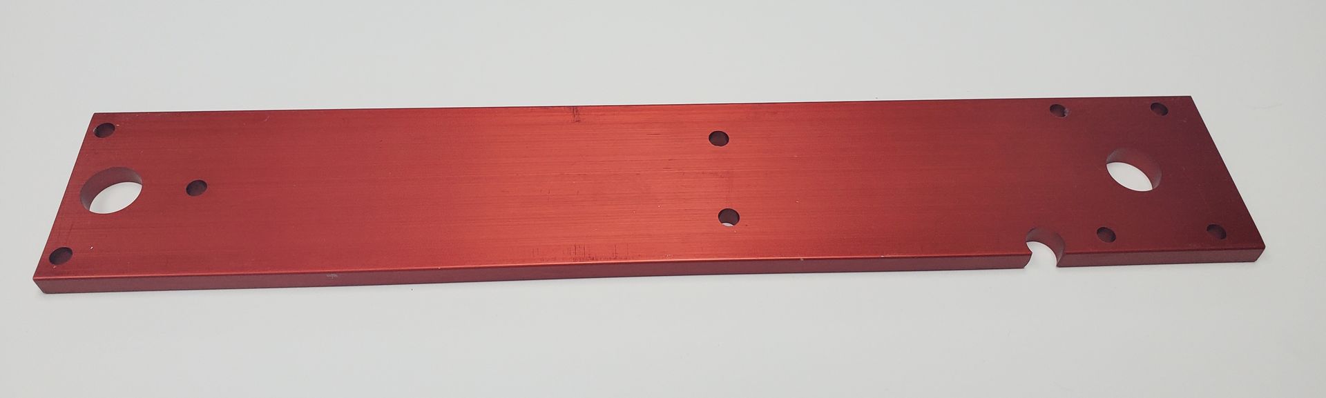 A red rectangular metal plate with several pre-drilled holes and a small notch on one side, lying on a white surface.