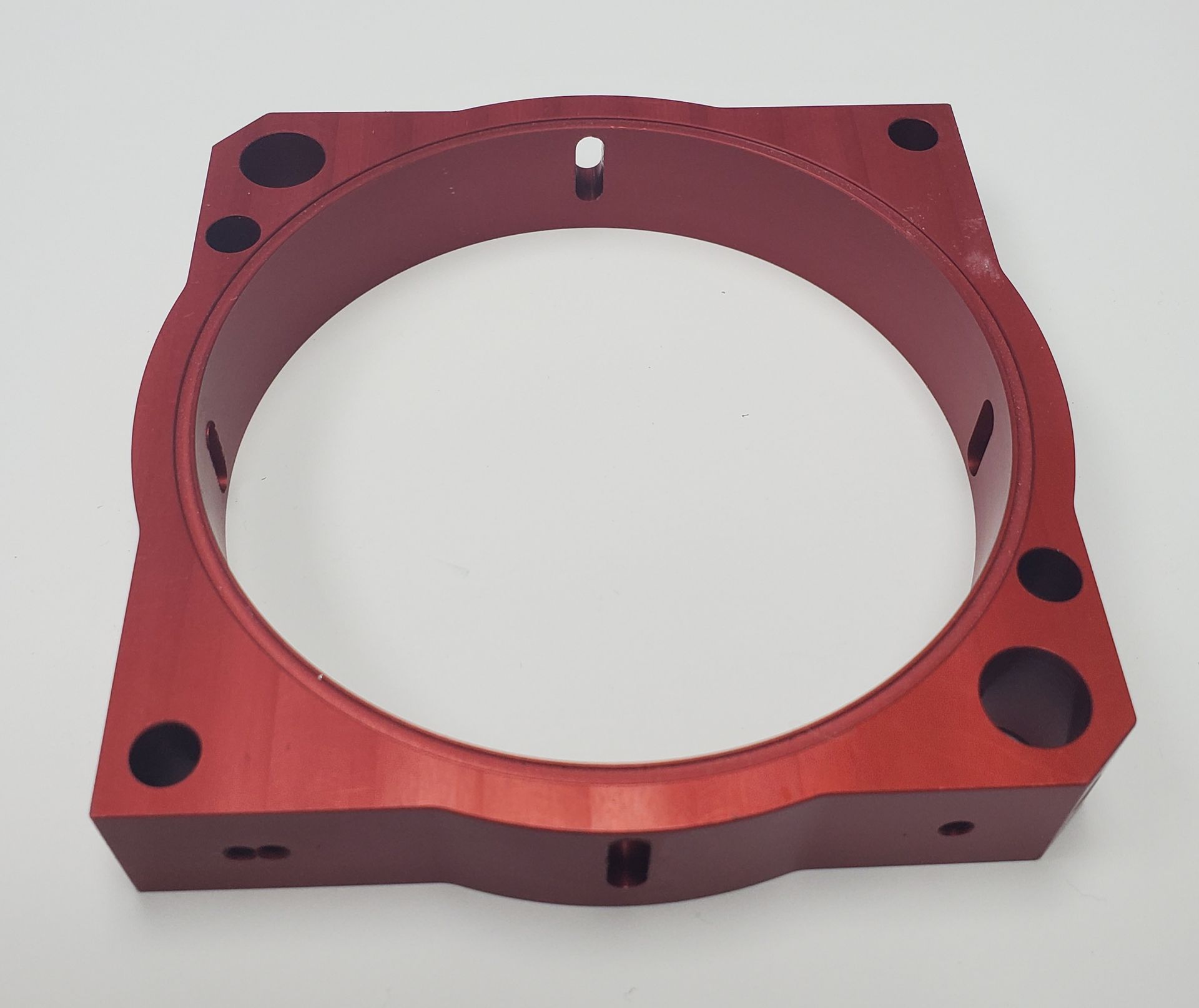 A red, square-shaped metal mechanical component with a large circular cutout in the center and various mounting holes.