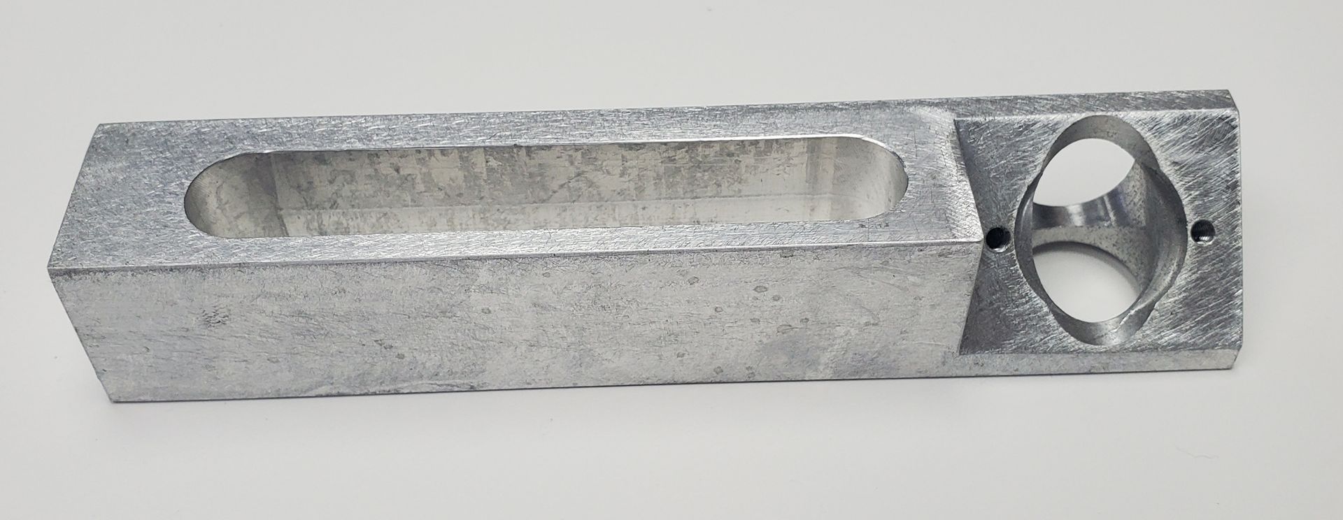 A rectangular, silver-colored metal piece featuring a long recessed slot and a circular opening with small side holes.