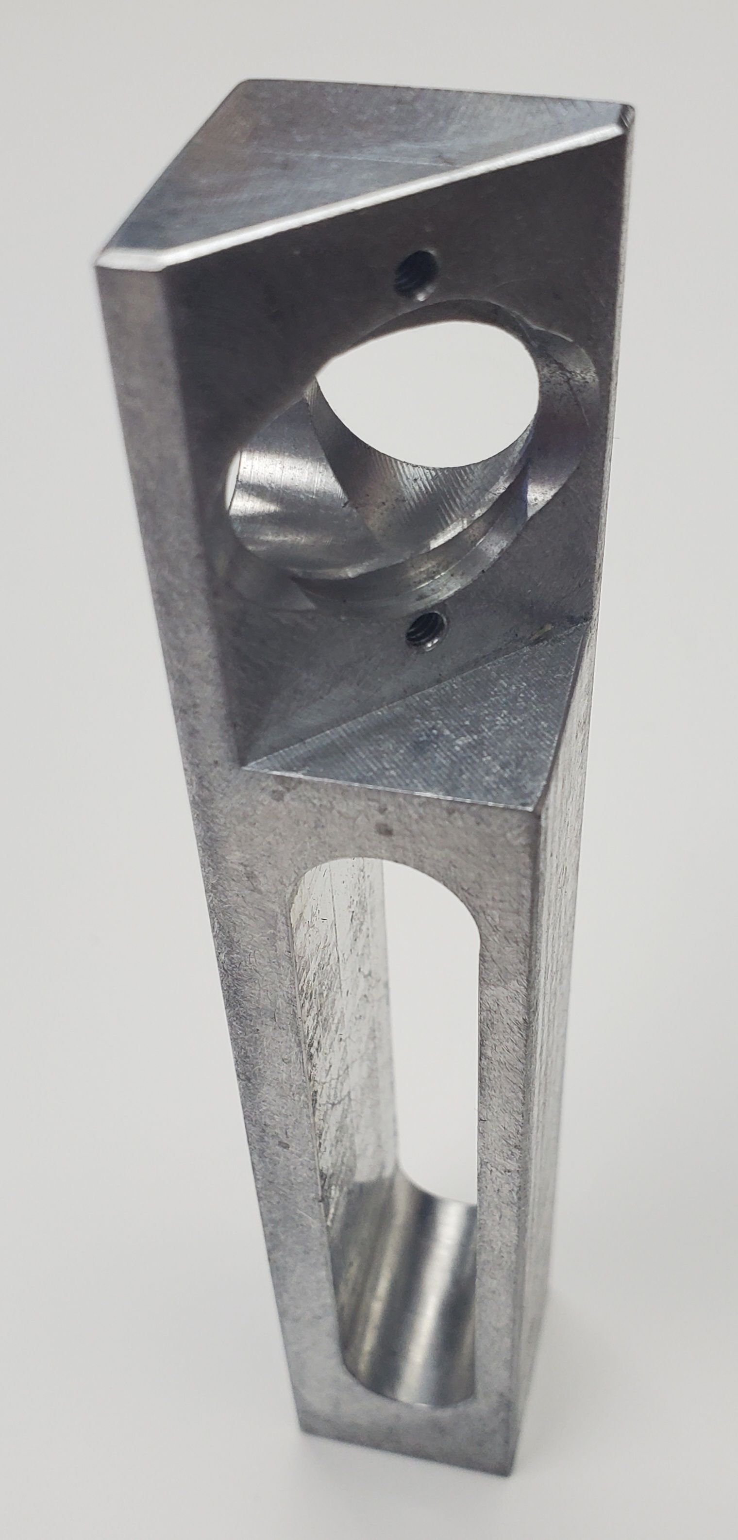 A tapered, metallic industrial part with a circular hole near the top and a vertical, elongated slot in the center.
