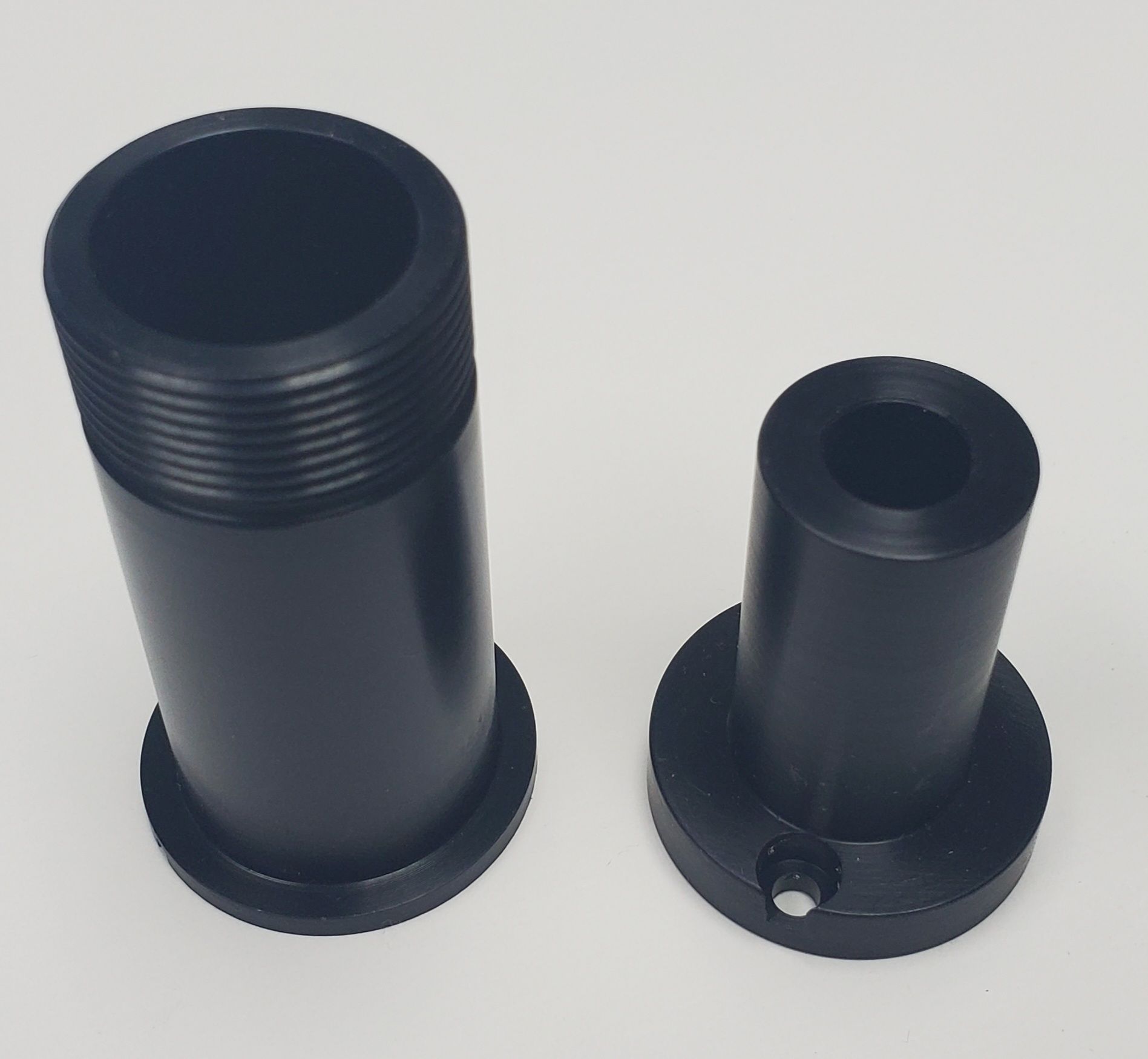 Two black, cylindrical mechanical components with flanges, one with exterior threading, sit side-by-side on a white surface.