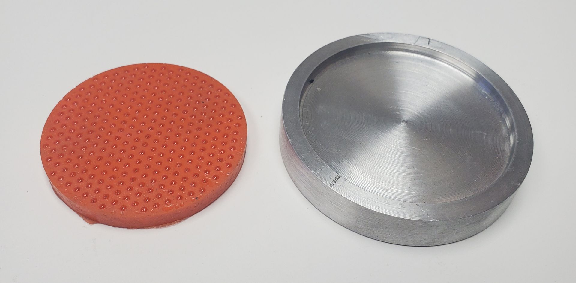 A round, orange, textured silicone pad next to a circular, silver metal container lid on a white surface.
