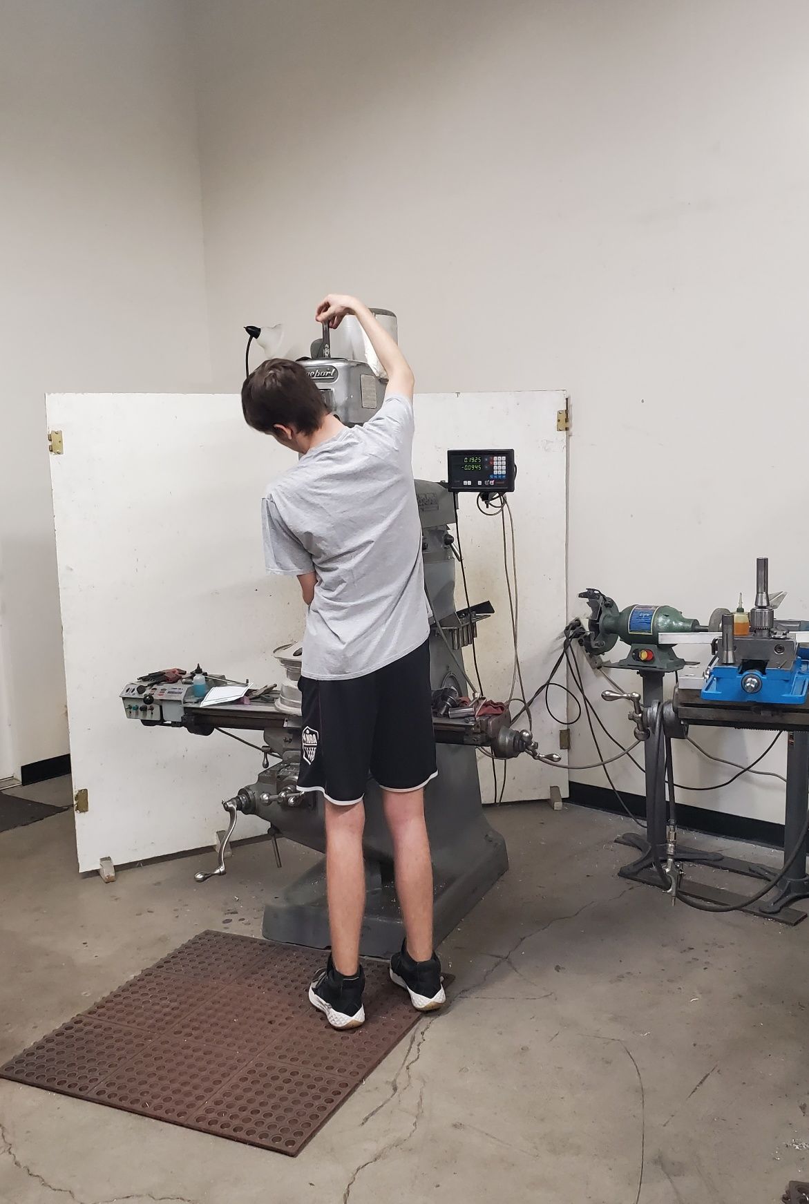 A person in a gray t-shirt and shorts stands at a metal milling machine in a workshop, adjusting the machine's head.
