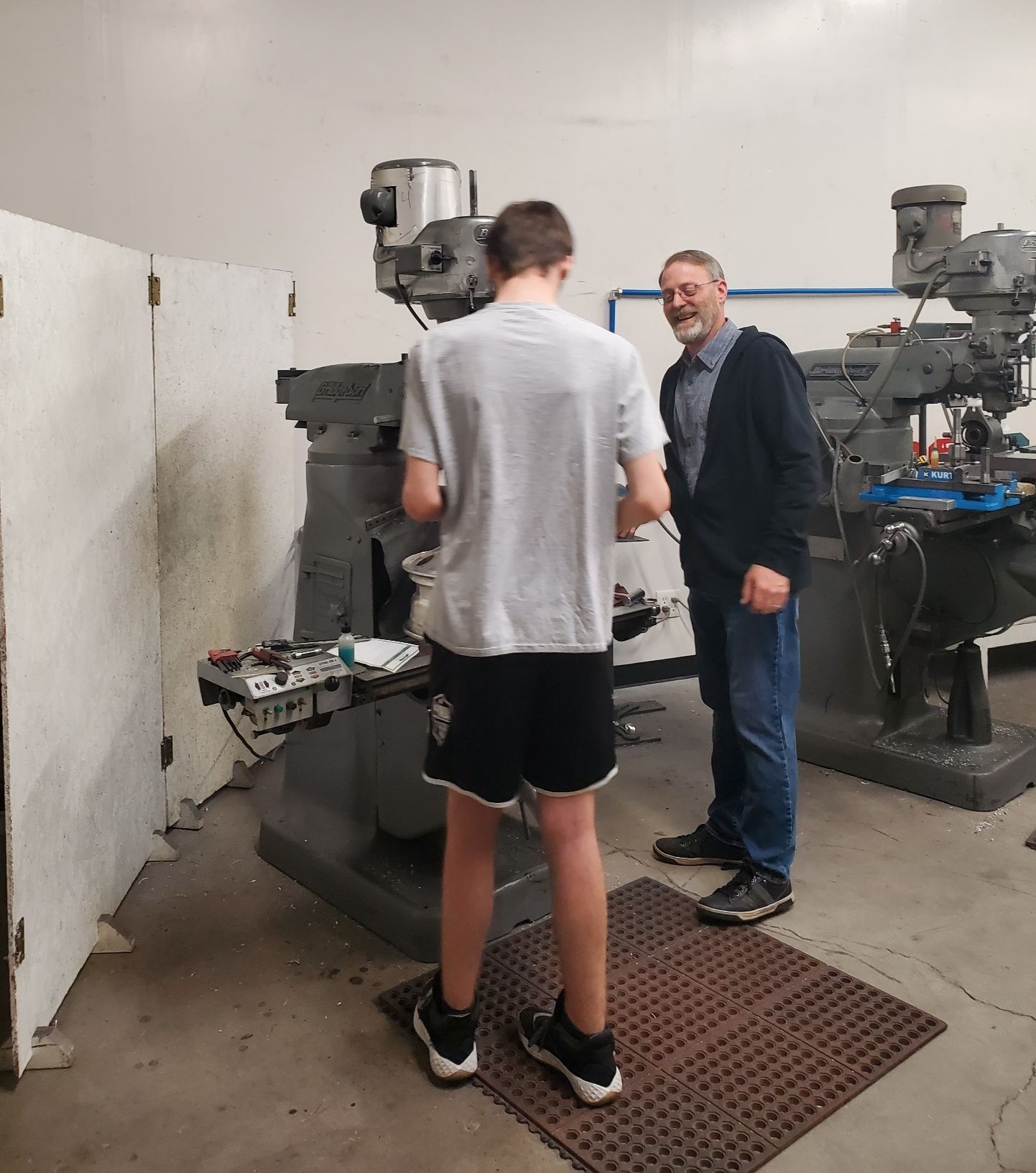 A person operates a milling machine while an instructor looks on in a workshop.