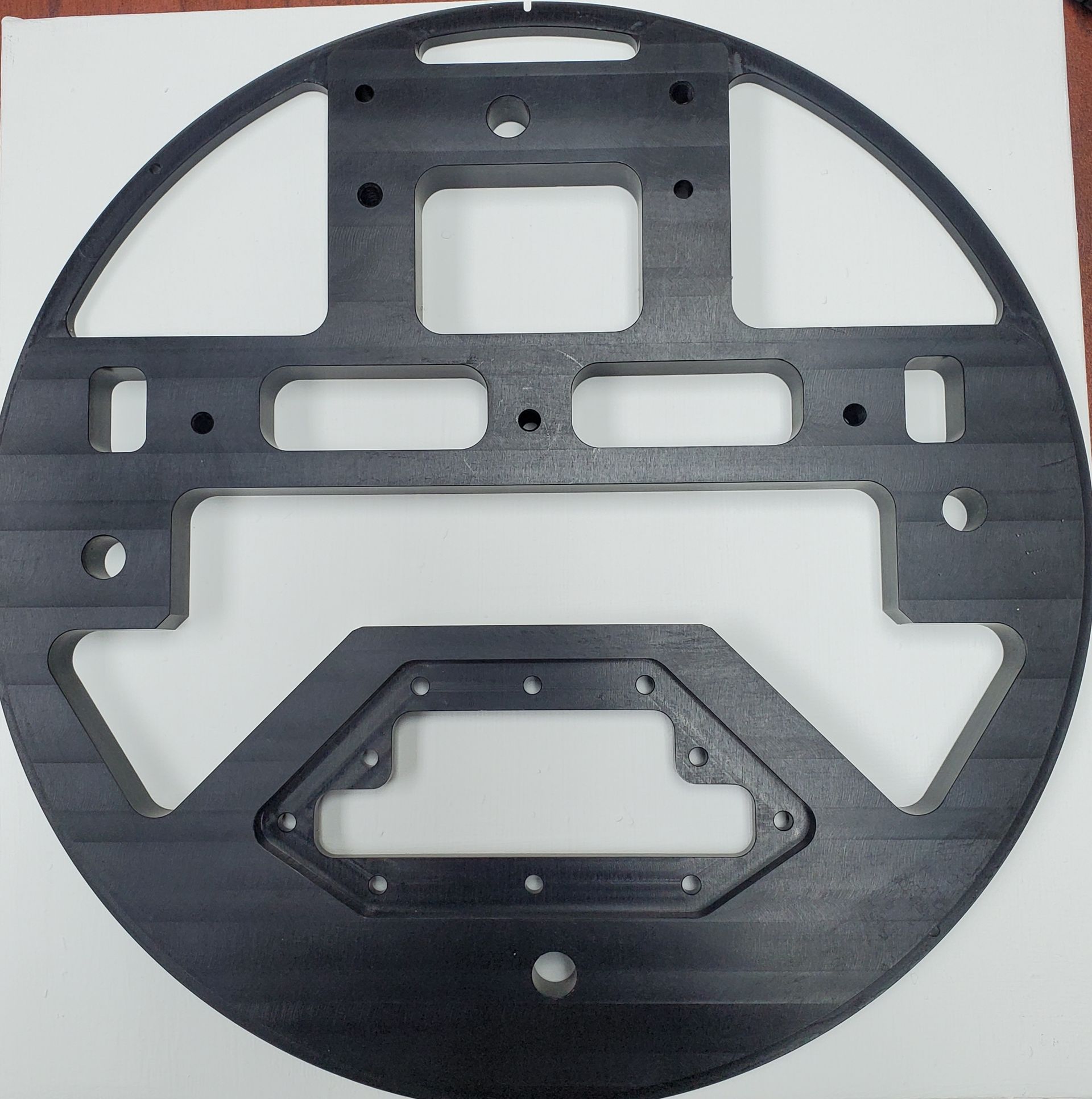 A black, circular 3D-printed mounting plate with various geometric cutouts and mounting holes for mechanical assembly.