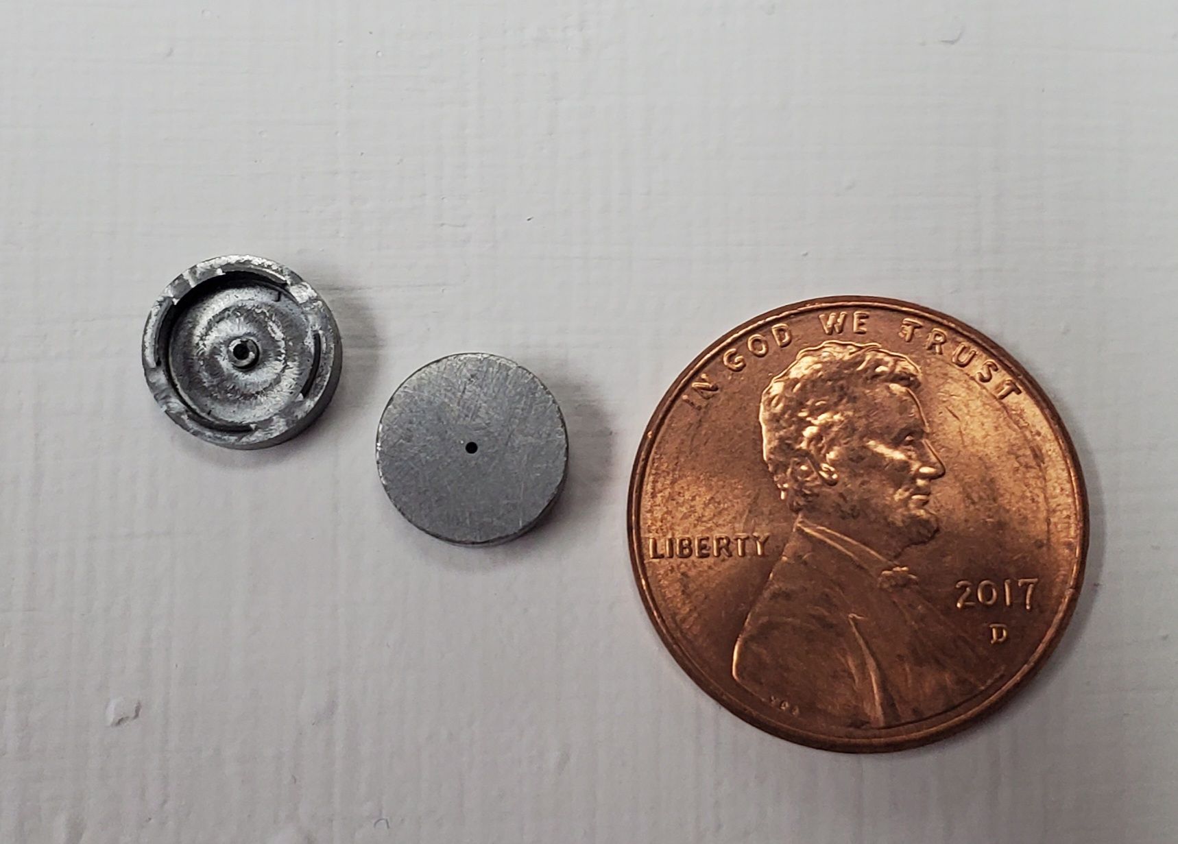 Two small, round, grey metal components next to a copper penny for scale on a white surface.