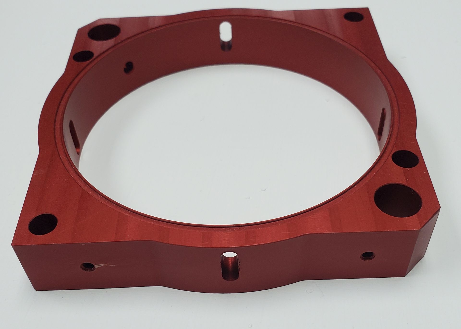 A red, square-shaped machined metal ring with circular mounting holes and slots, set against a plain white background.
